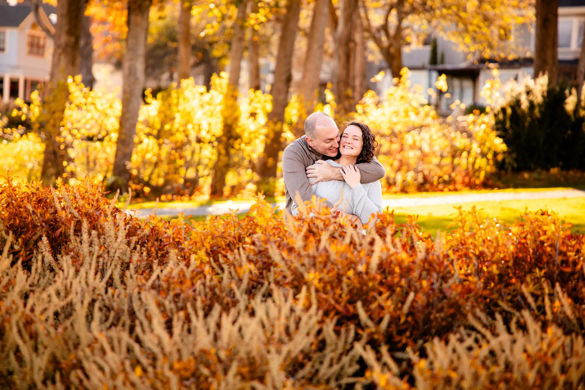 A man hugs a smiling woman while standing behind yellow bushes during engagement portraits at the Bar Harbor Inn and Spa in Maine.