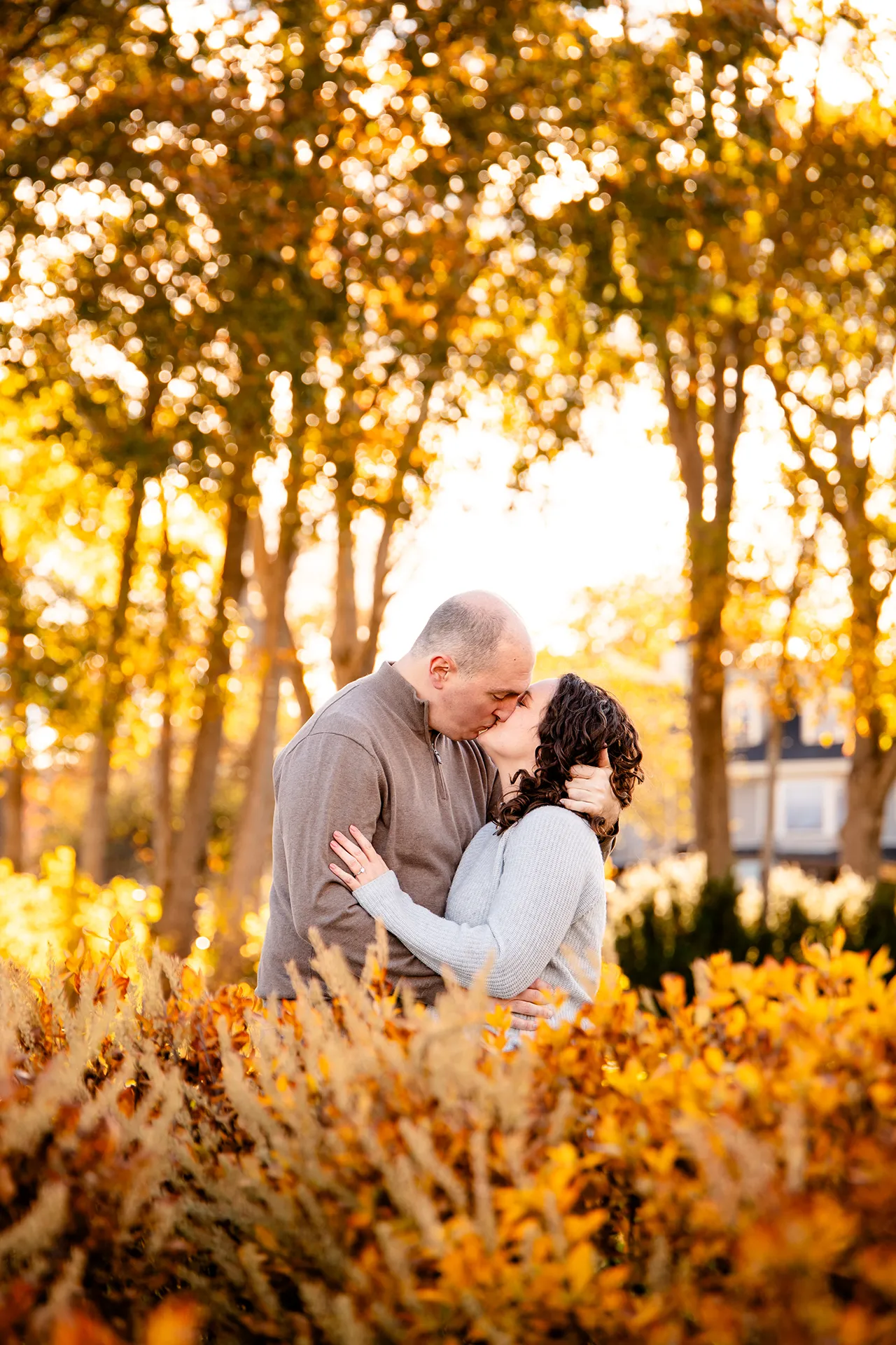 A man and woman kiss while standing behind yellow bushes during an engagement session at the Bar Harbor Inn and Spa near Acadia National Park.