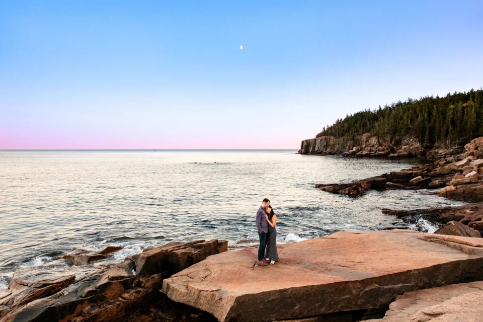 A newly engaged couple snuggle together as the sun sets during an engagement session at Otter Cliff in Acadia National Park, Maine.
