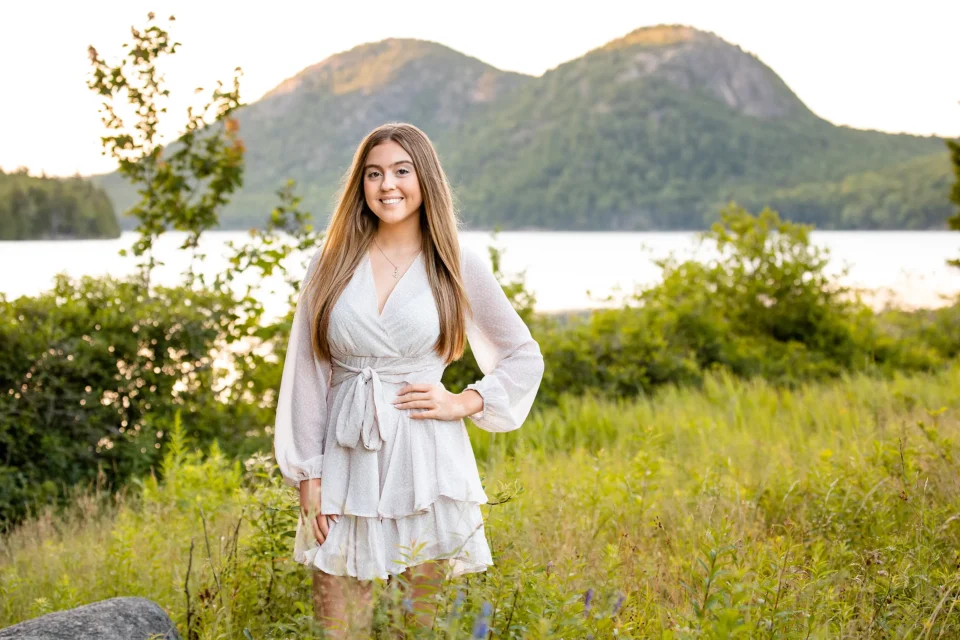 A girl smiles and poses in a field during a senior portrait session at Jordan Pond in Acadia National Park near Bar Harbor, Maine.