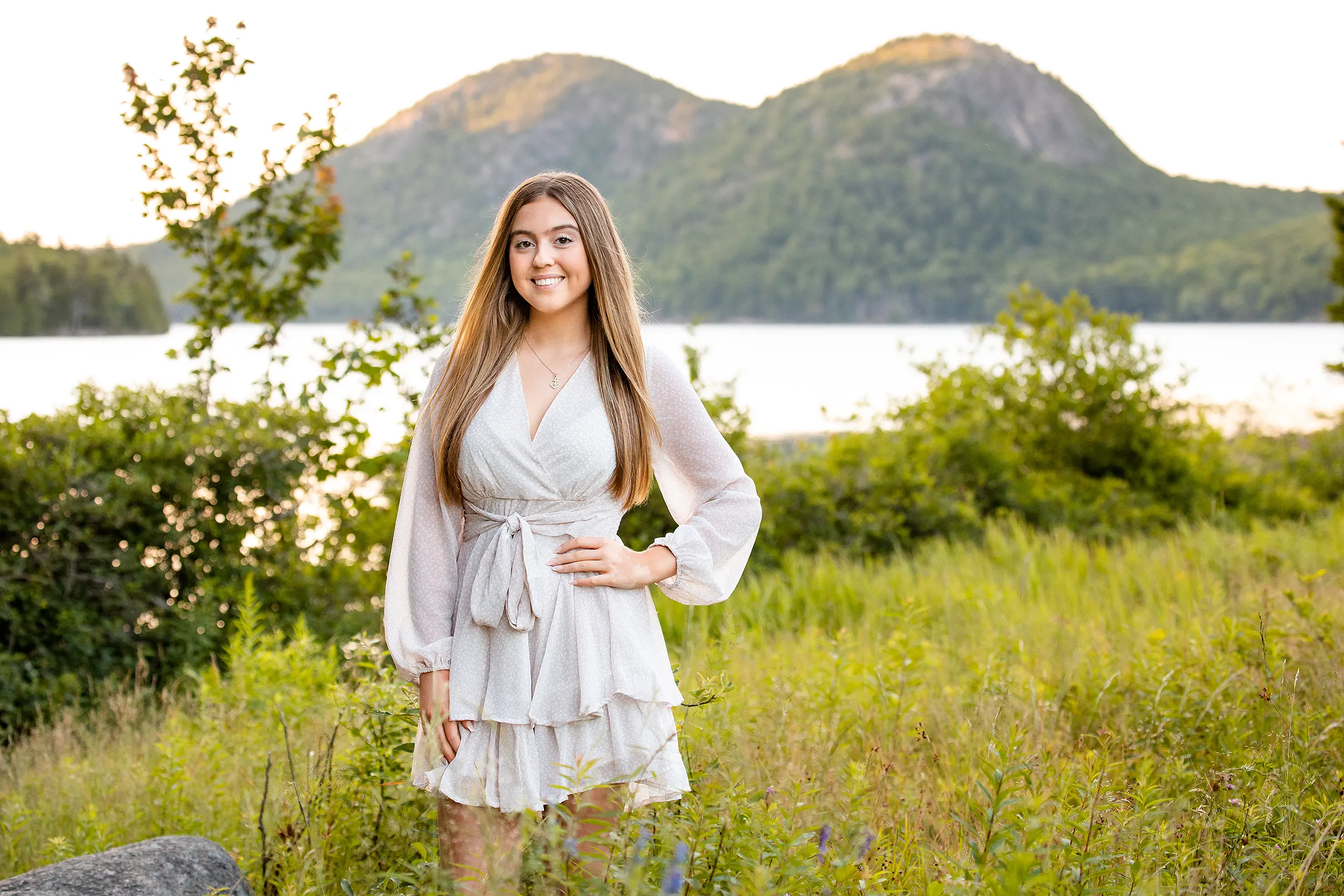 A girl smiles and poses in a field during a senior portrait session at Jordan Pond in Acadia National Park near Bar Harbor, Maine.