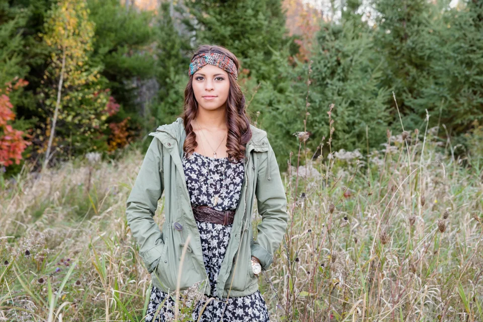 A girl smiles and poses in a field during a senior portrait session at the Bangor City Forest in Maine.