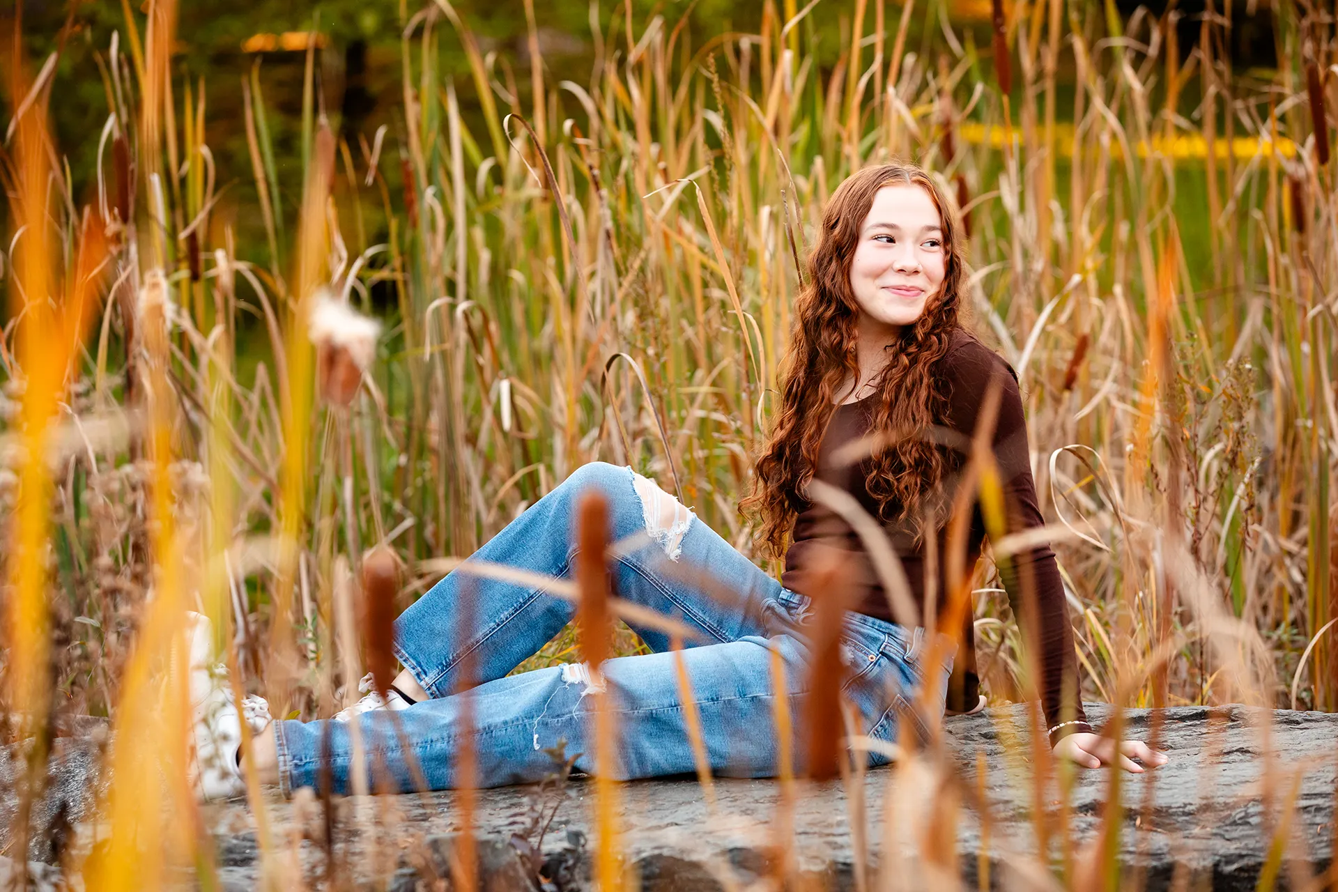 A high school senior sits amidst tall yellow grass and cat o' nine tails at the LIttlefield Ornamentals Garden at the University of Maine.