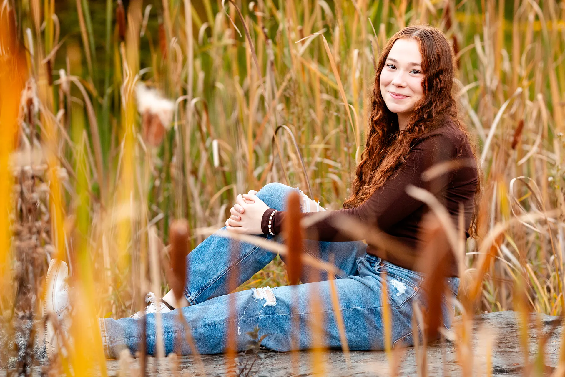 A high school senior sits and poses amidst tall yellow grass and cat o' nine tails at the LIttlefield Ornamentals Garden at the University of Maine.