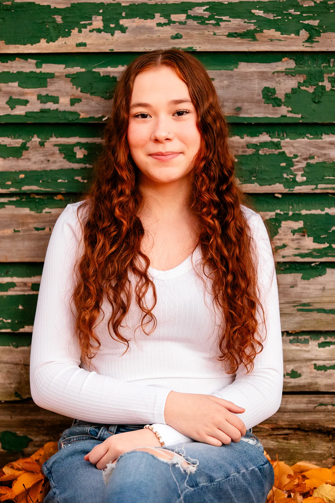 A high school senior sits in fall leaves leaning against a green wooden building during a portrait session at the LIttlefield Ornamentals Garden.