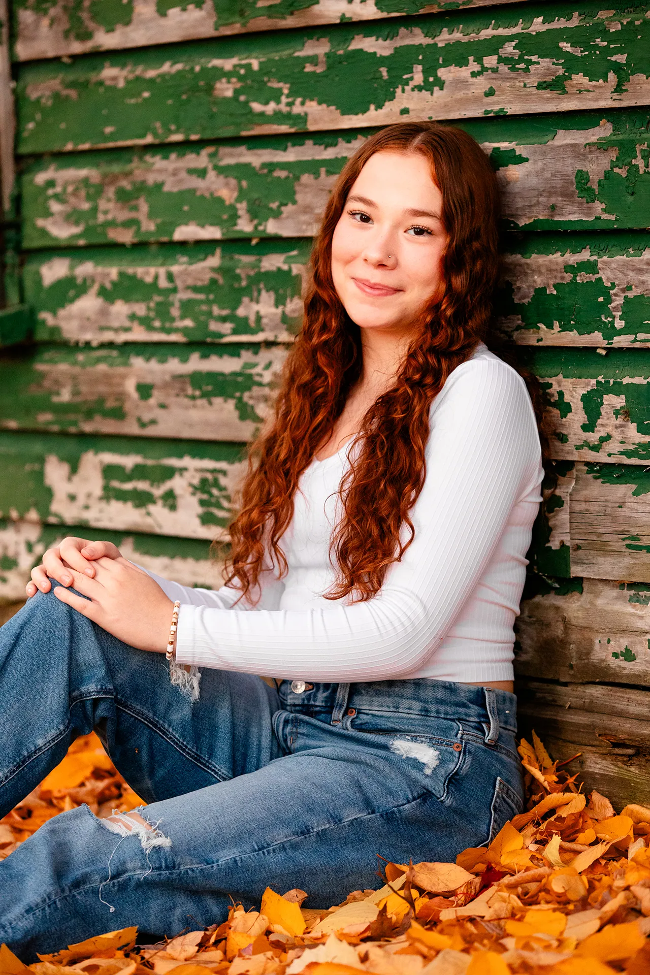 A high school senior sits in fall leaves leaning against a green wooden building during a portrait session at the University of Maine.
