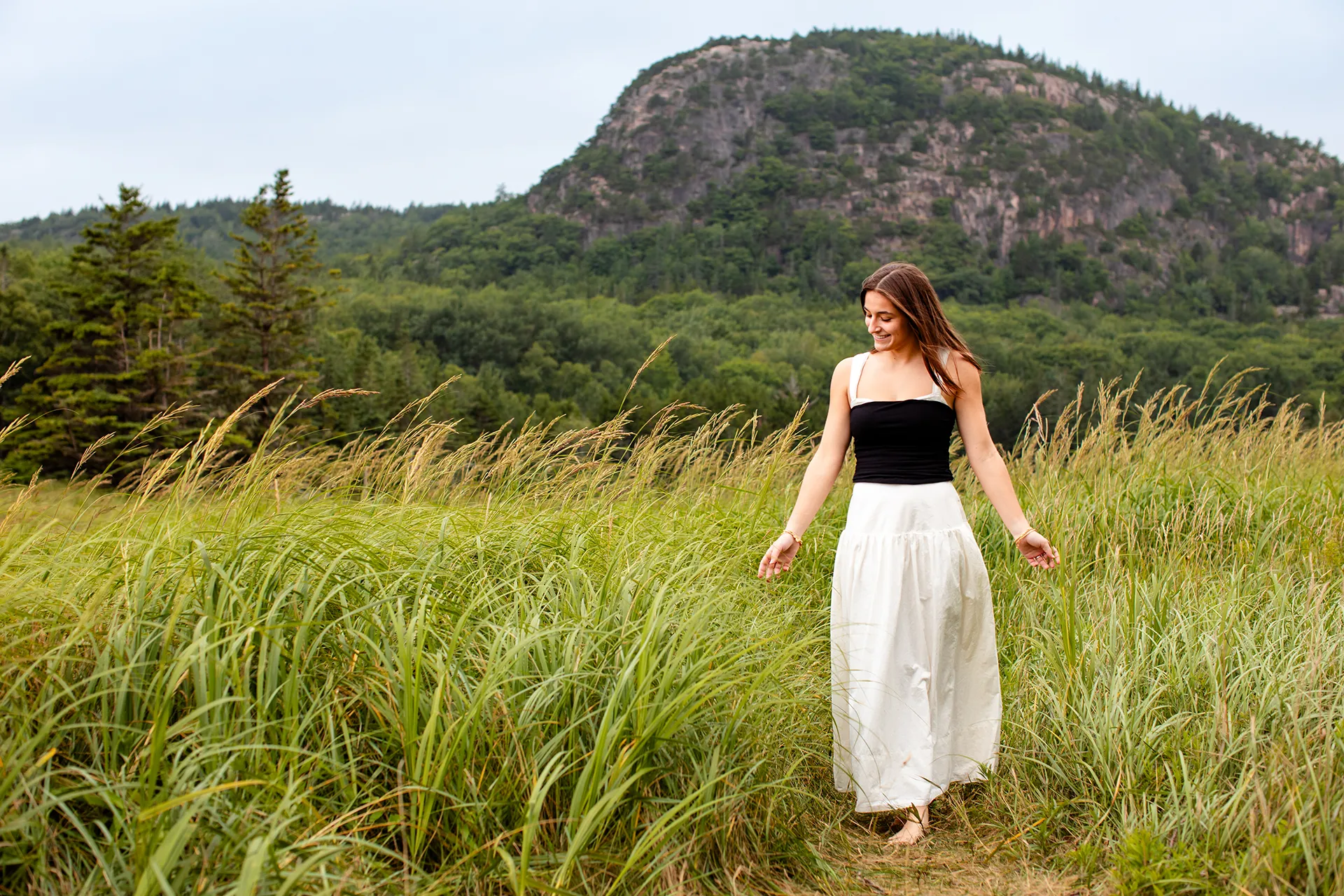 A girl walks through tall grass in front of Beehive Mountain during a senior portrait session at Sand Beach in Acadia National Park.