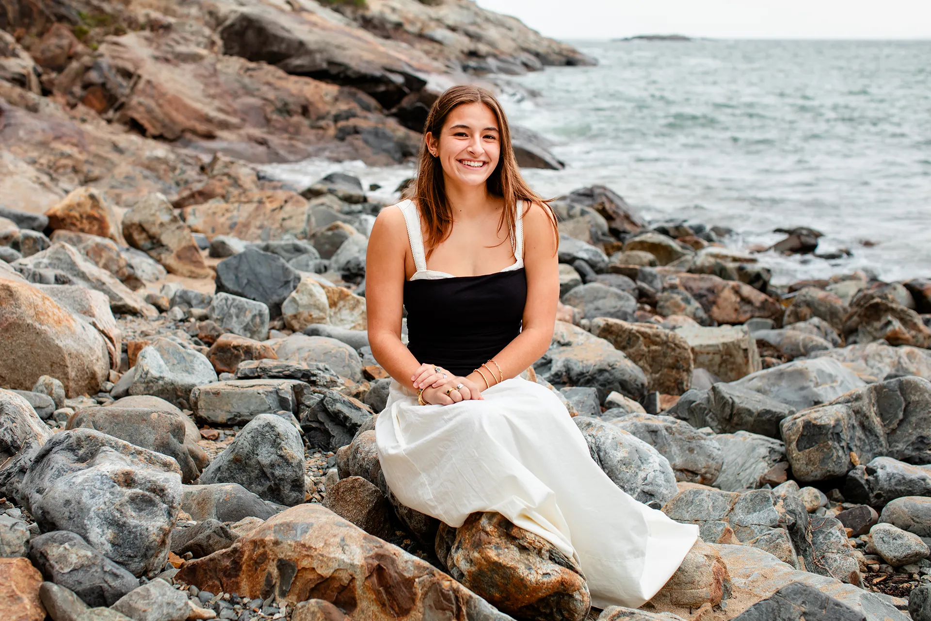 A girl laughs as she sits on some rocks during a senior portrait session at Sand Beach in Acadia National Park.