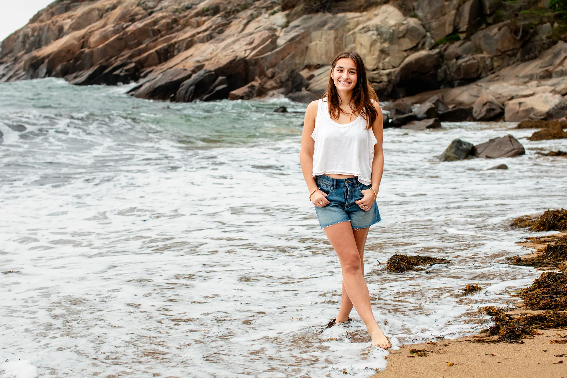 A girl smiles as she poses in the ocean during a senior portrait session at Sand Beach in Acadia National Park.