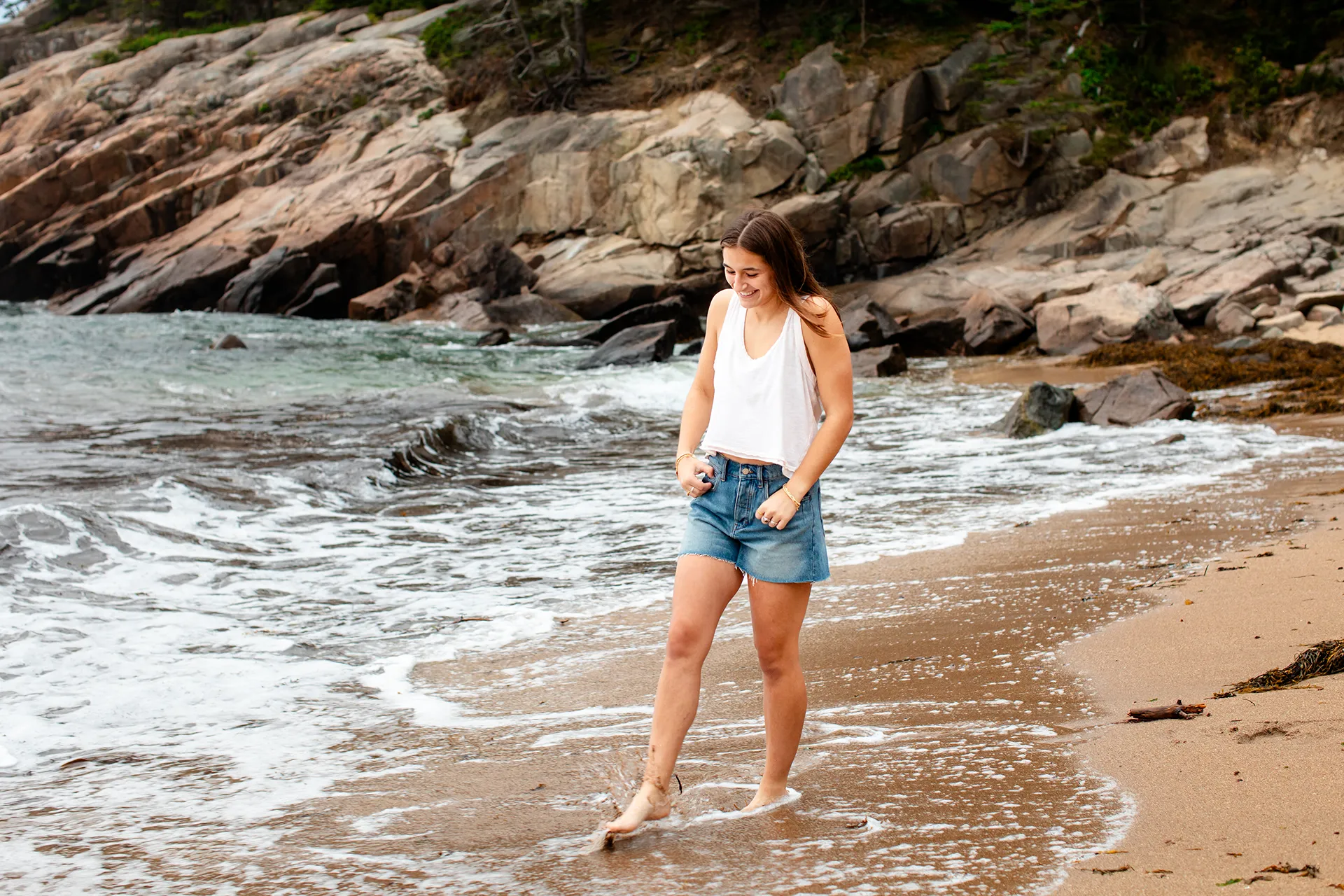 A girl smiles as she walks through the ocean during a senior portrait session at Sand Beach in Acadia National Park.