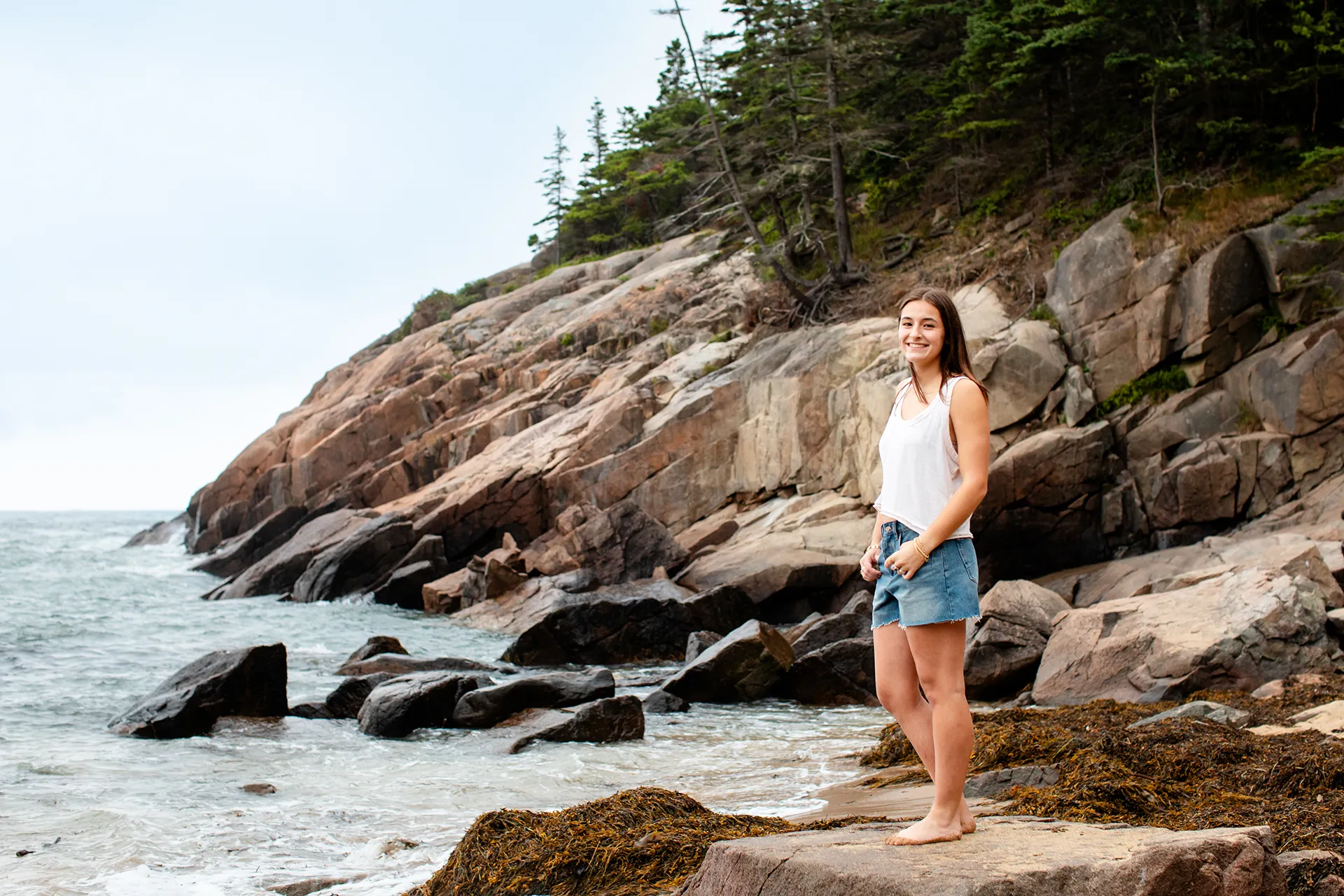 A girl poses on a rock in front of the ocean during a high school senior portrait session at Sand Beach in Acadia National Park.