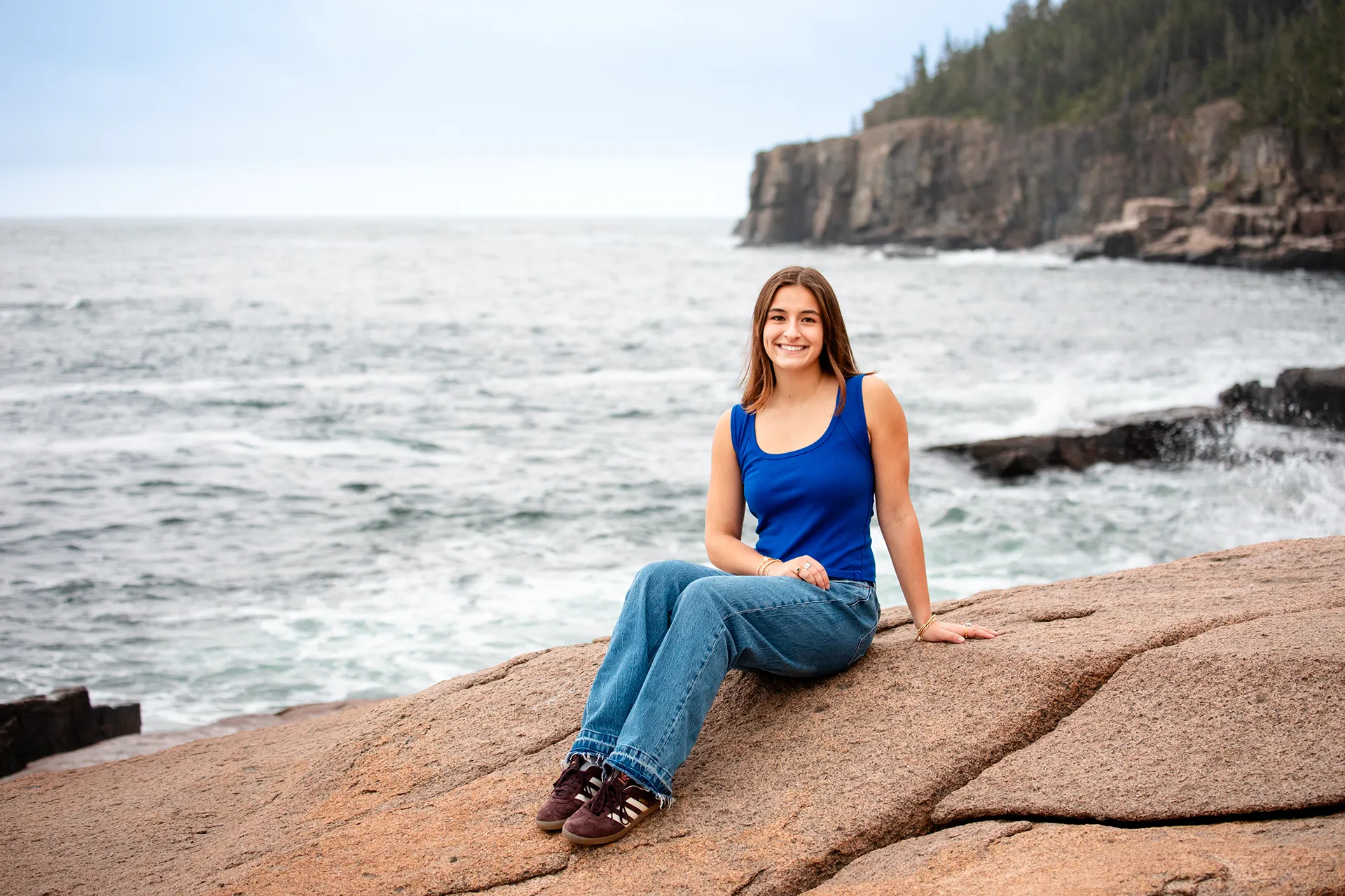 A high school senior smiles as she sits by the ocean during a high school senior portrait session near the Otter Cliff in Acadia National Park.