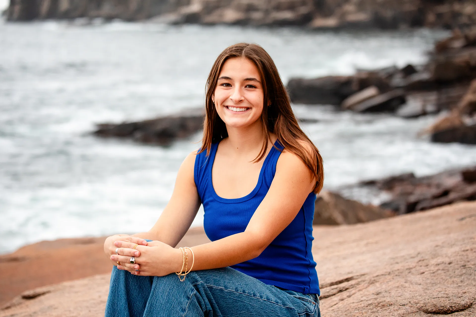 A girl smiles as she sits on a rock during a high school senior portrait session near the Ocean Path in Acadia National Park.