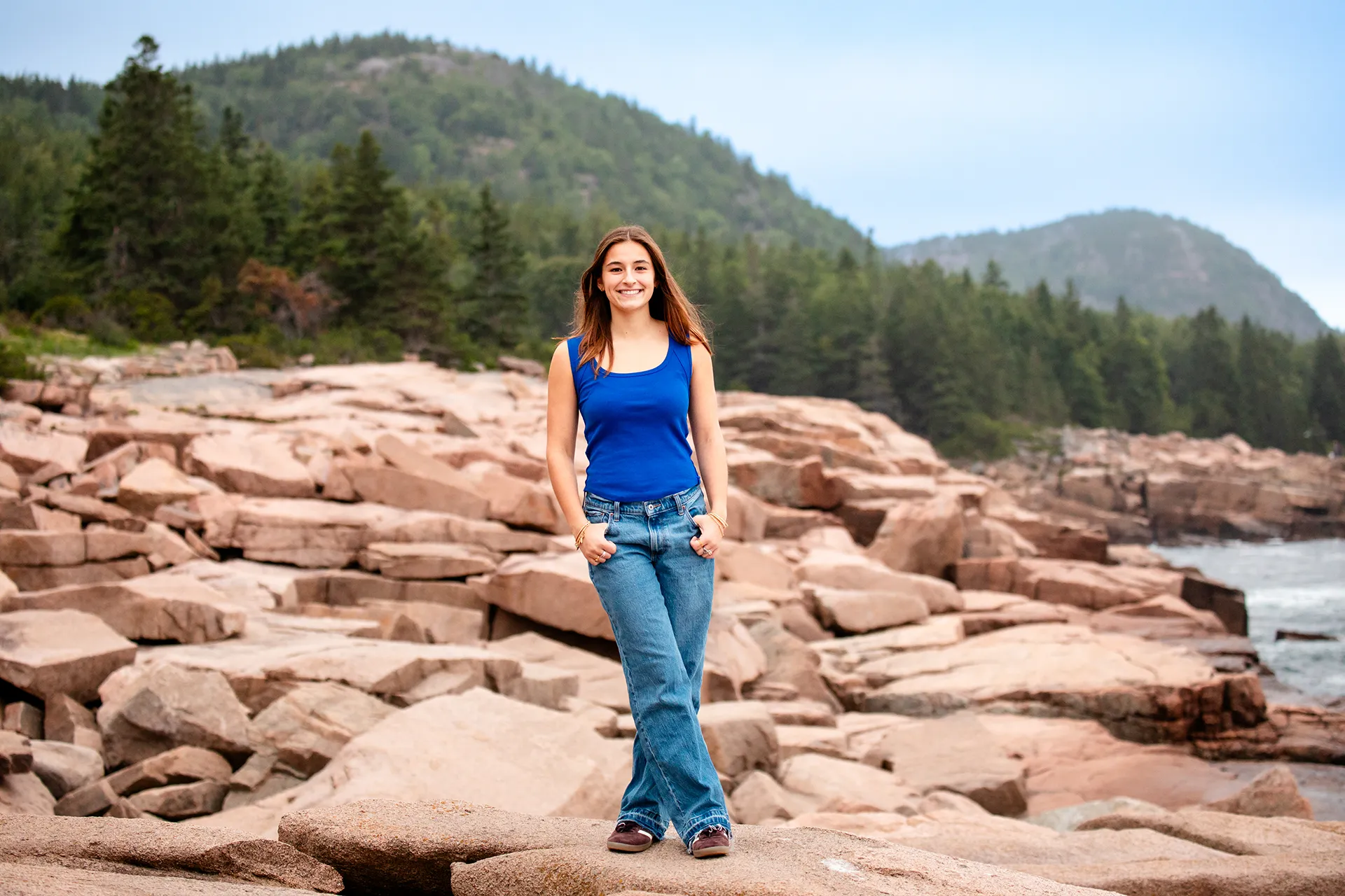 A girl smiles and poses in front of Beehive Mountain during a high school senior portrait session near the Ocean Path in Acadia National Park.