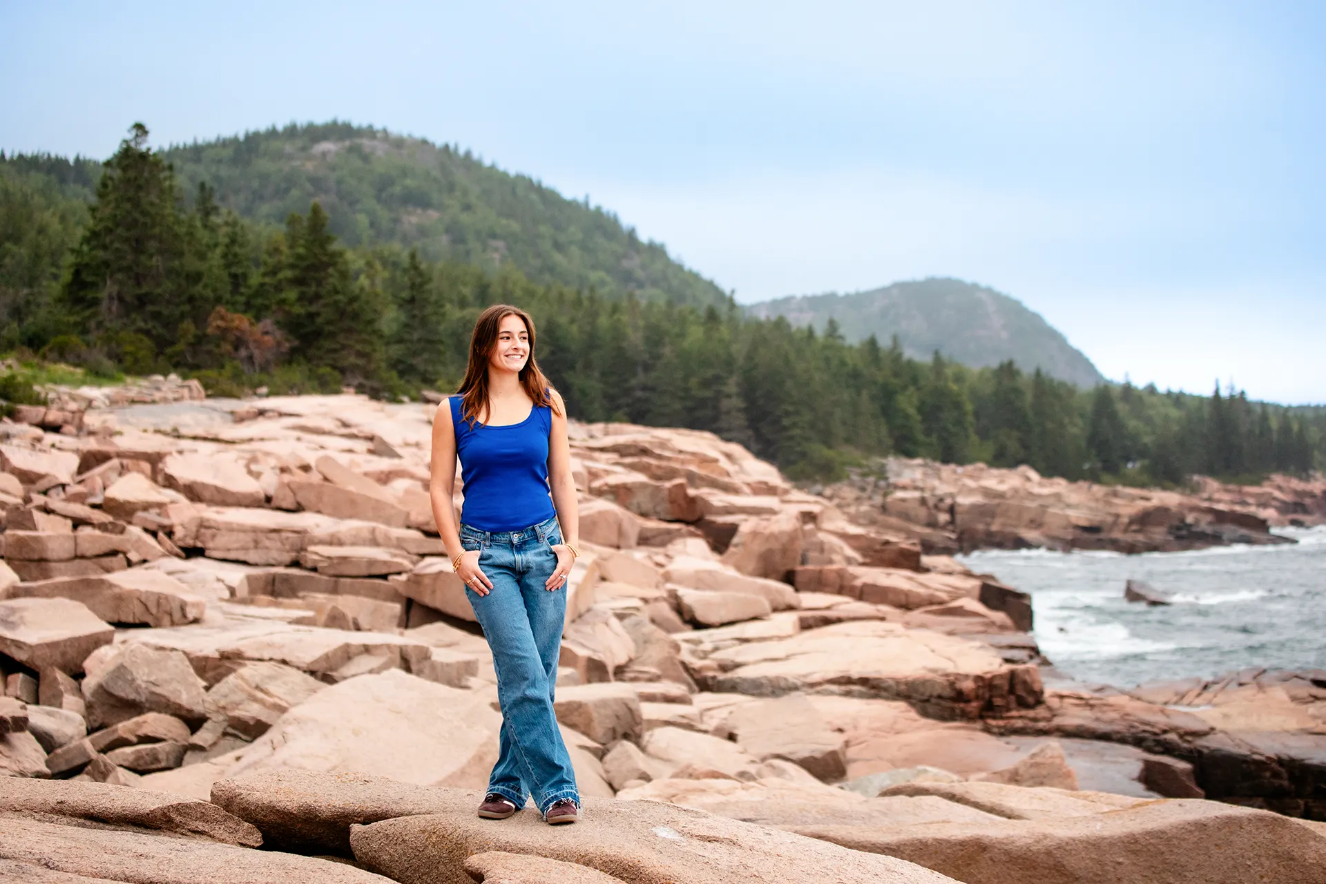 A high school senior smiles and poses on a rocky coastline in front of Beehive Mountain near the Ocean Path in Acadia National Park.