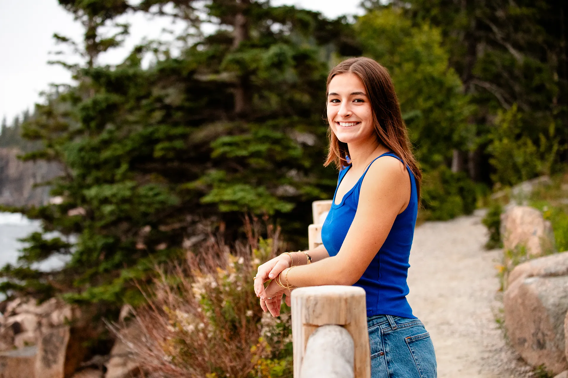 A girl smiles as she rests her on arms on a fence during a high school senior portrait session on Ocean Path in Acadia National Park, Maine.