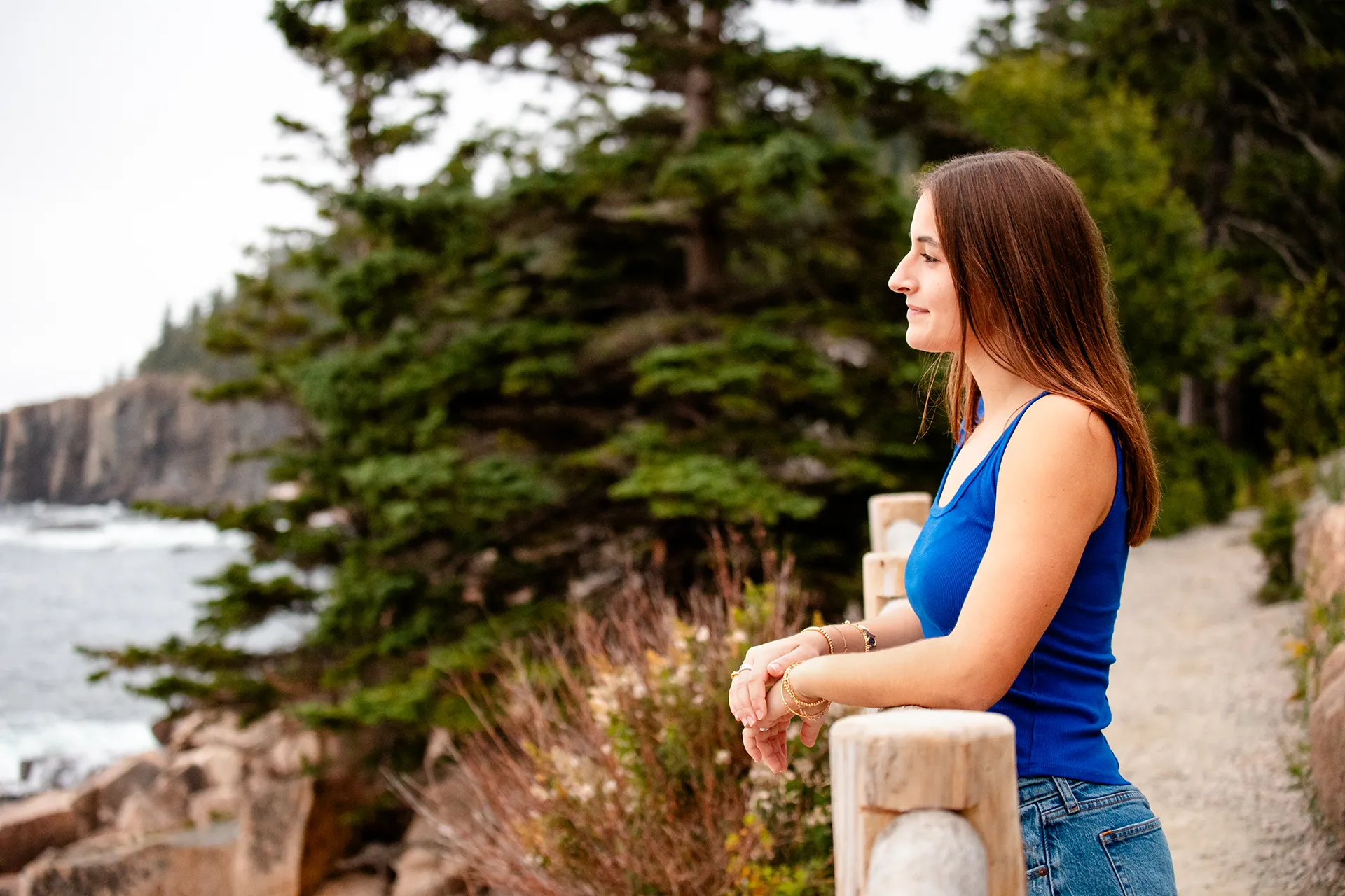 A girl looks off towards the ocean as she rests her on arms on a fence during a senior portrait session on Ocean Path in Acadia National Park, Maine.