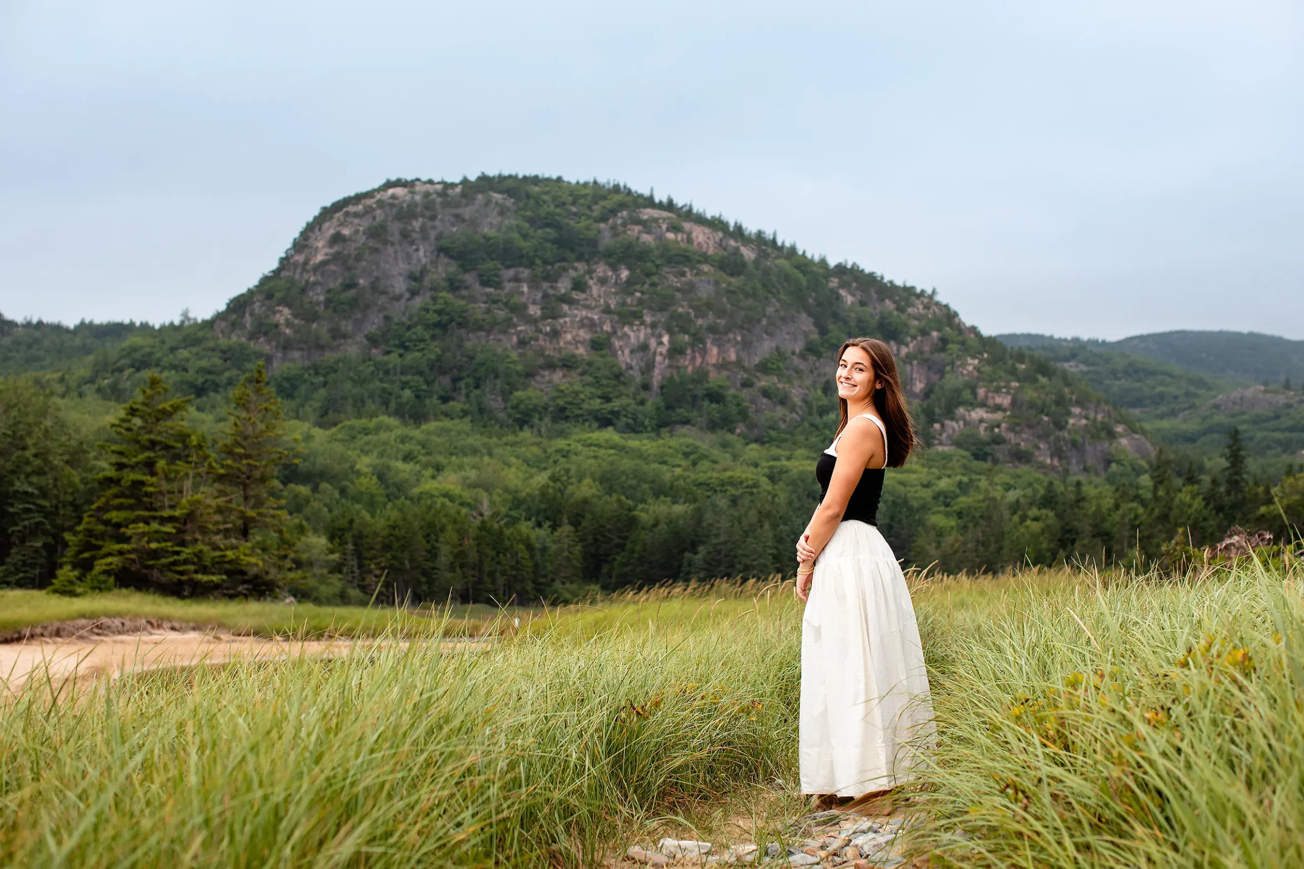 A girl poses in tall grass in front of Beehive Mountain during a senior portrait session at Sand Beach in Acadia National Park.