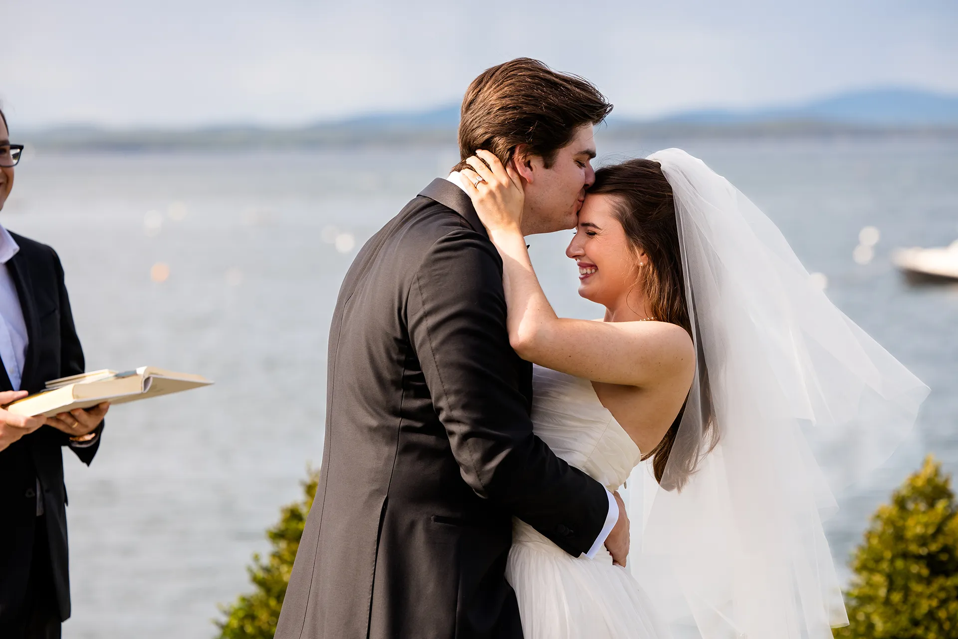 A groom kisses a bride on the top of the head while she smiles during a wedding ceremony at the Bar Harbor Inn and Spa in Maine.