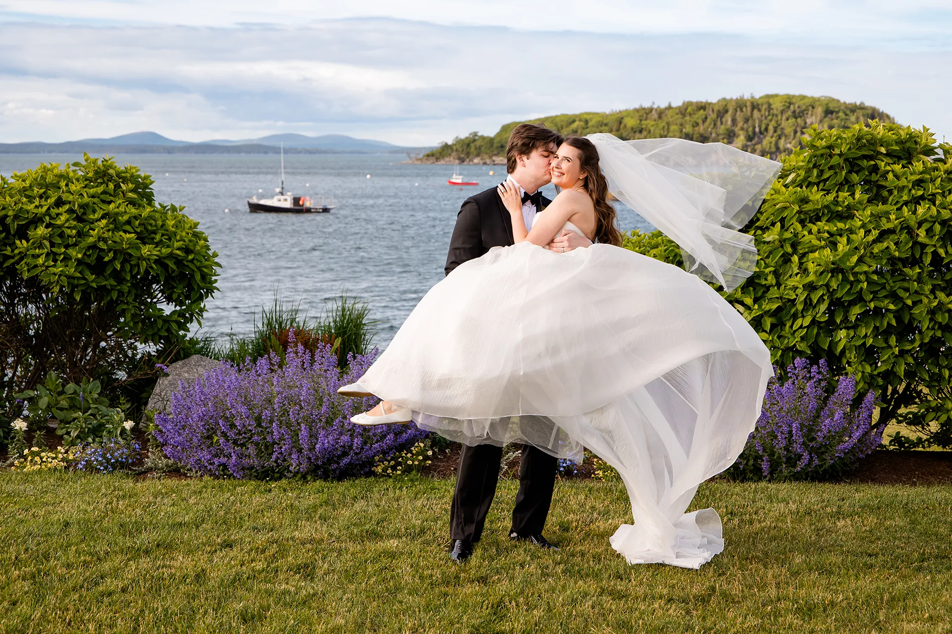 A groom holds a bride while he kisses her cheek and her dress blows in the wind during wedding portraits at the Bar Harbor Inn and Spa in Maine.