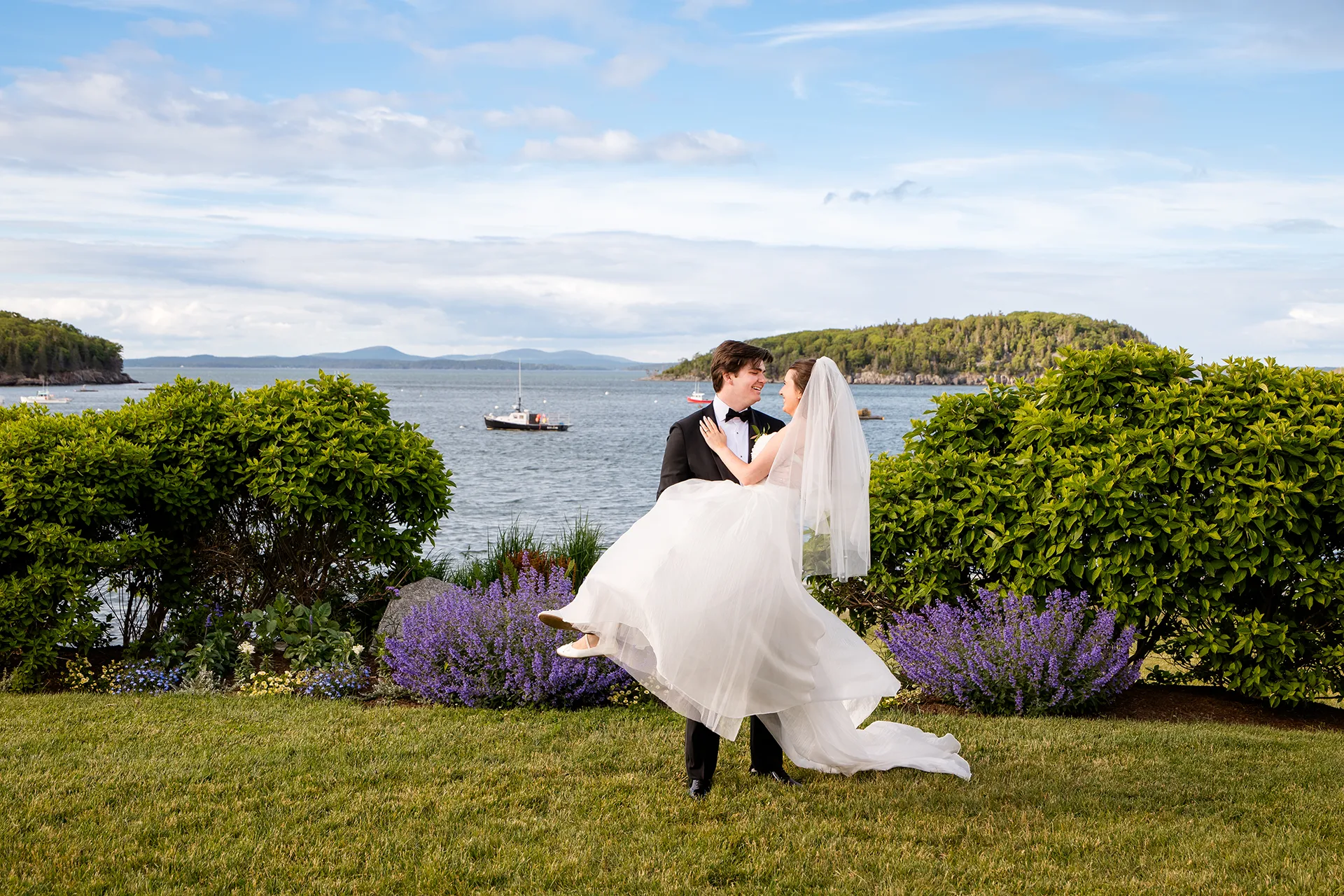 A groom smiles at a bride while he holds her during wedding portraits in front of Frenchman Bay on the property of the Bar Harbor Inn and Spa in Maine.