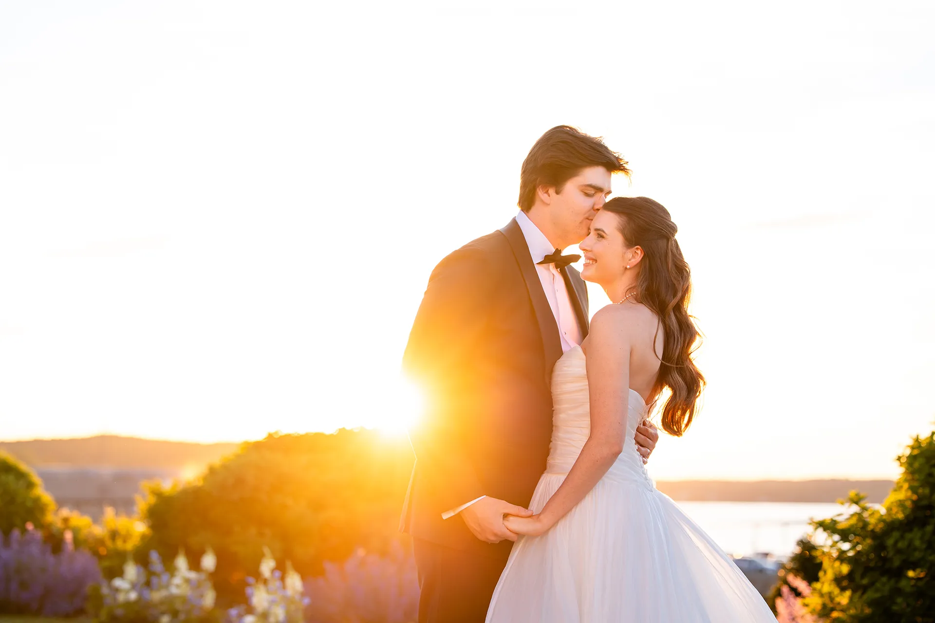 A groom kisses a smiling bride on the forehead as the sunsets during their wedding portraits at the Bar Harbor Inn and Spa in Maine.