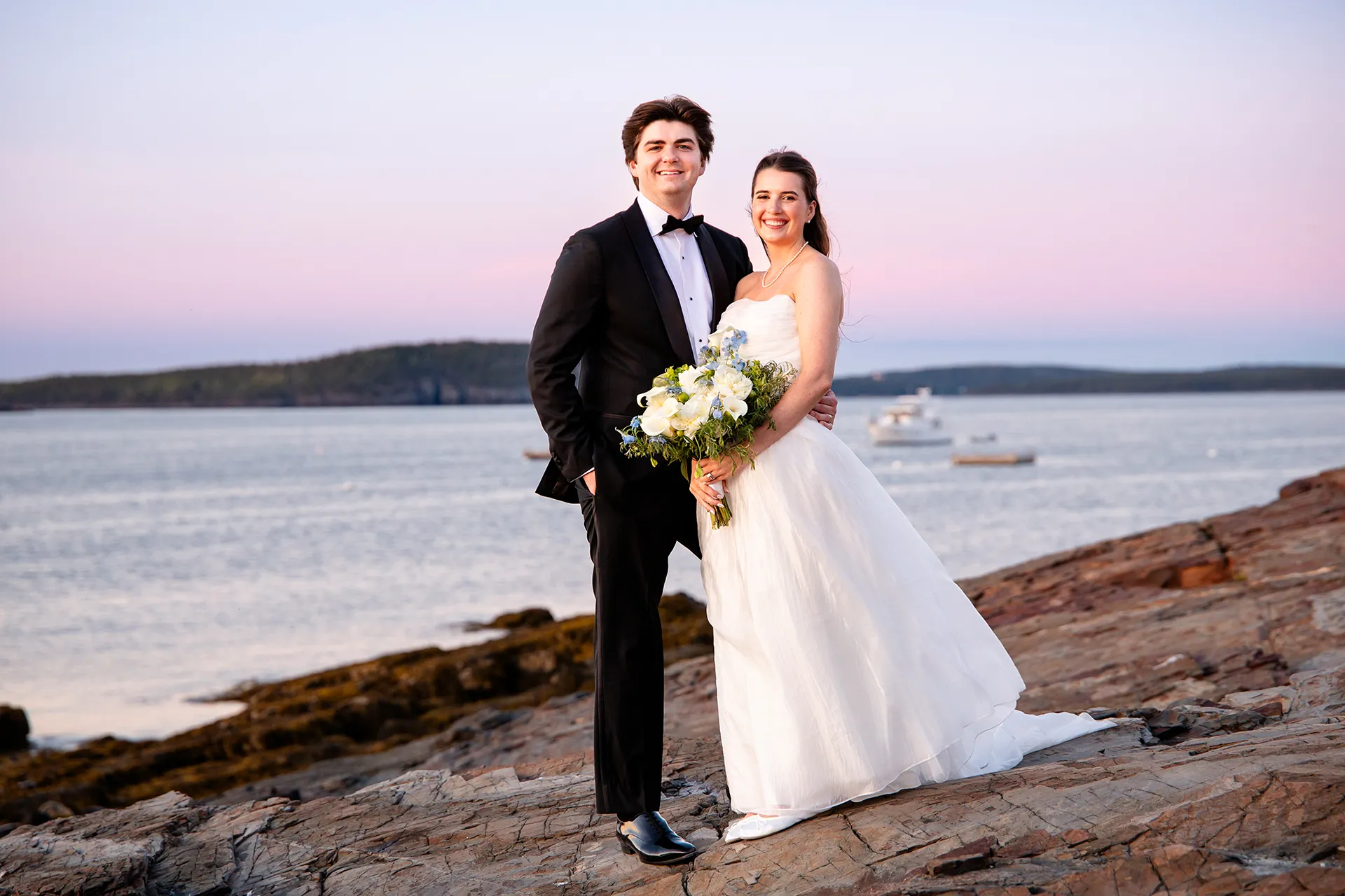 A newly married couple smile and pose in front of Frenchman Bay during their sunset wedding portraits at the Bar Harbor Inn and Spa in Maine.