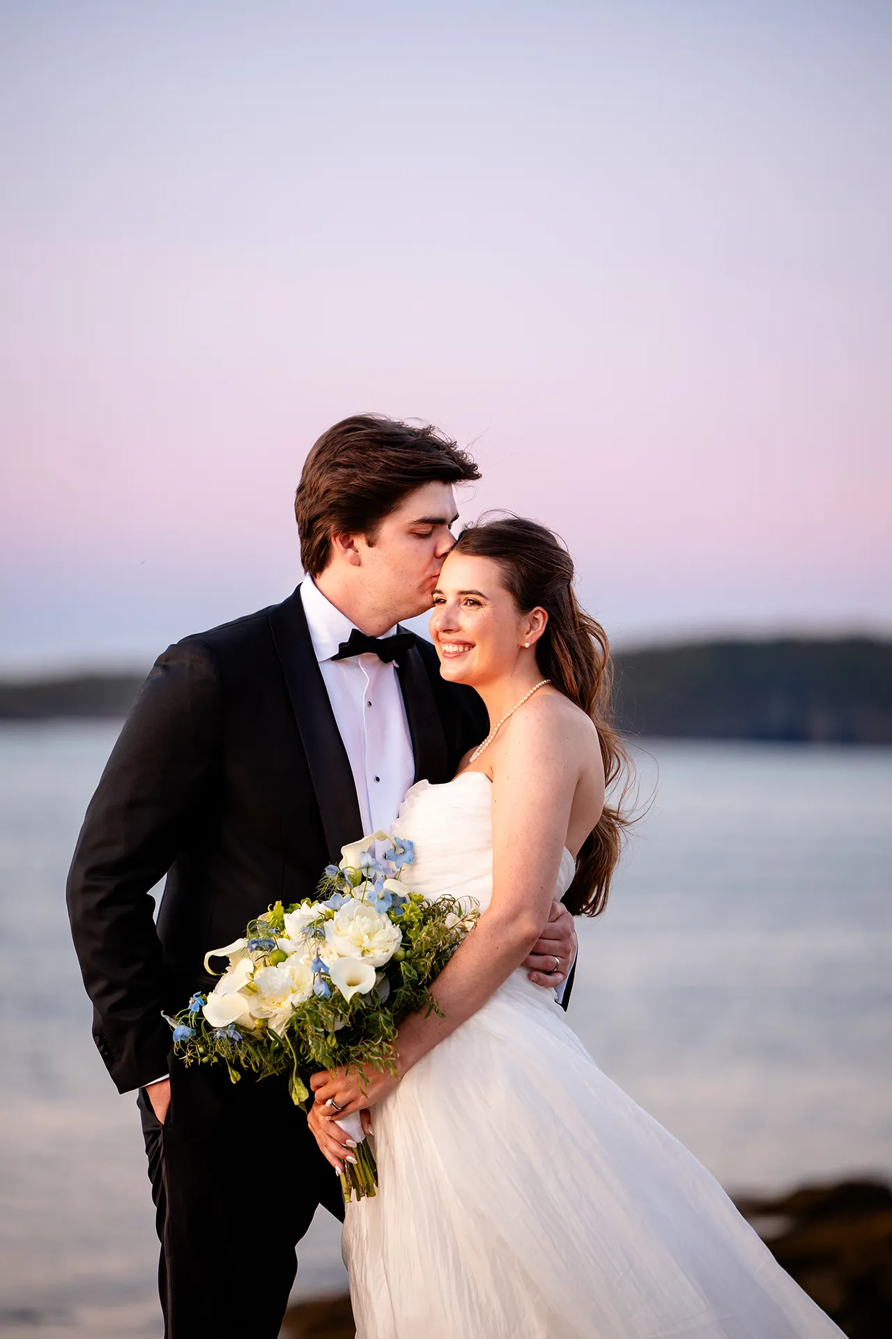 A bride smiles as groom kisses her forehead during wedding portraits at the Bar Harbor Inn and Spa near Acadia National Park in Maine.