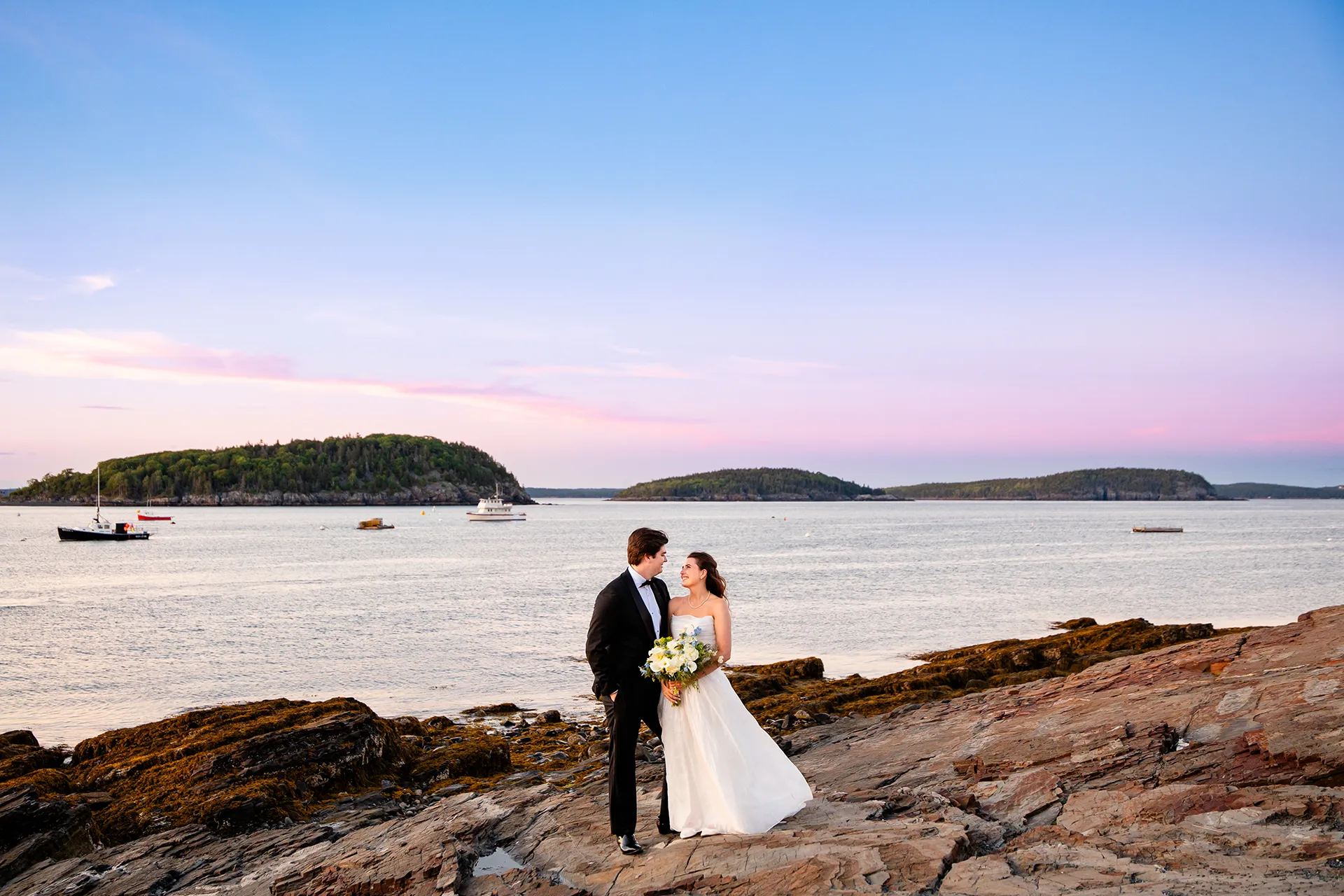 A bride and groom smile at each other while standing on rocks in front of Frenchman Bay during wedding portraits at the Bar Harbor Inn and Spa.