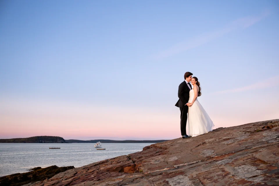 A bride smiles as groom kisses her forehead during their sunset wedding portraits at the Bar Harbor Inn and Spa in Maine.