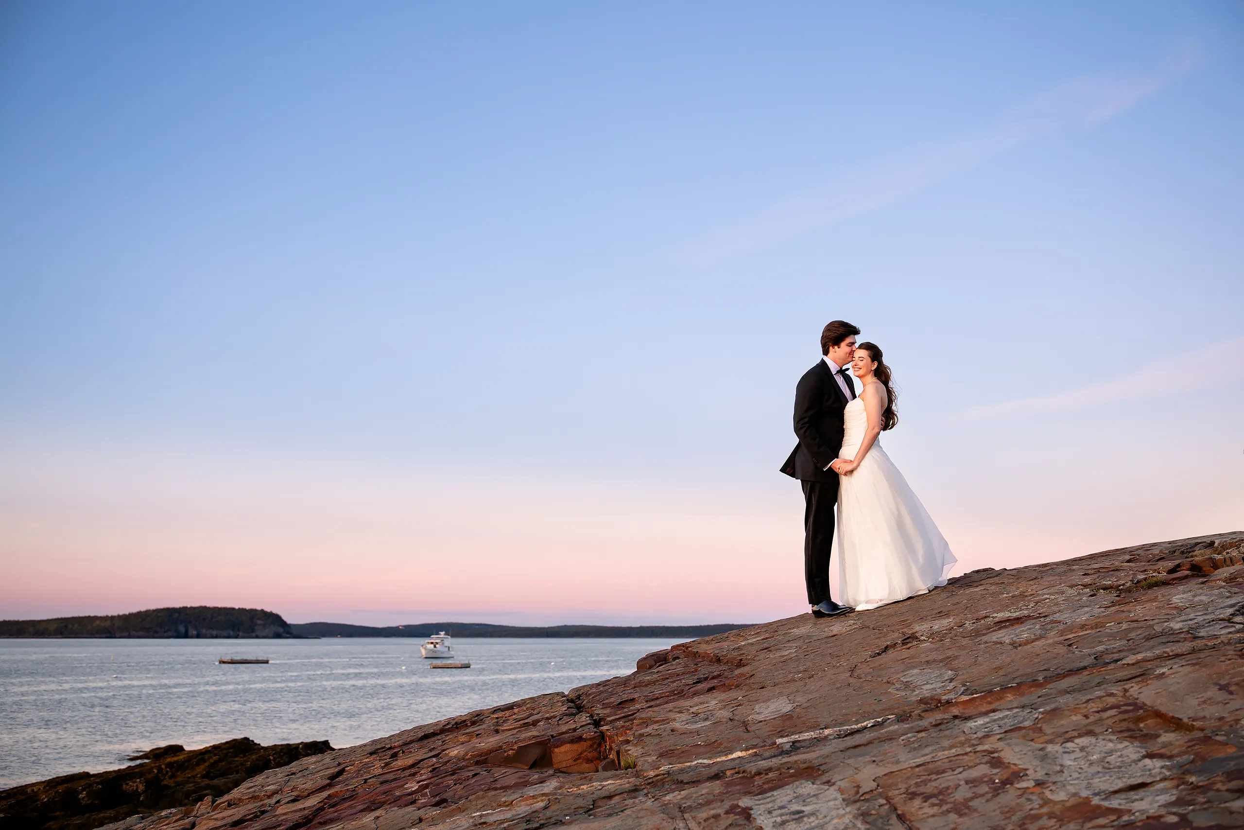 A bride smiles as groom kisses her forehead during their sunset wedding portraits at the Bar Harbor Inn and Spa in Maine.