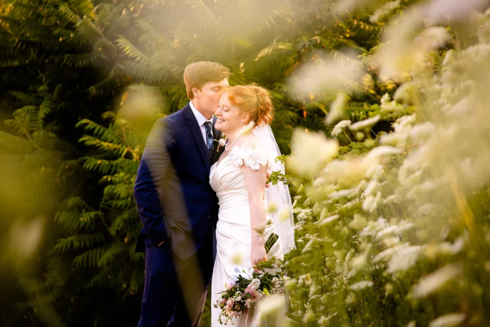 A groom kisses a bride on the cheek as they pose behind flowers during wedding portraits at the Hilton Garden Inn in Bangor, Maine.