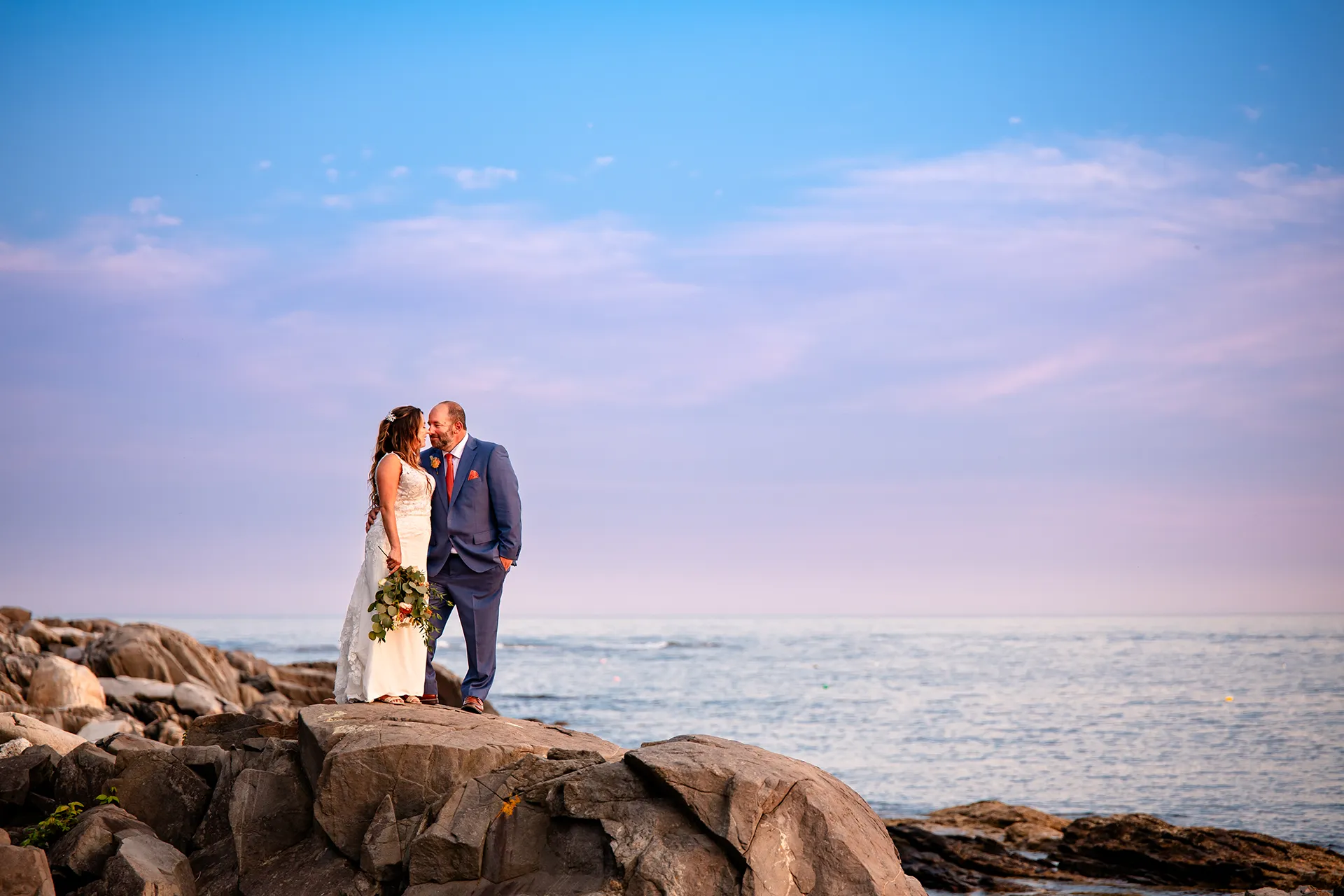 A bride and groom stand on a cliff at sunset as they snuggle together during wedding portraits at the Newagen Seaside Inn in Southport Maine.