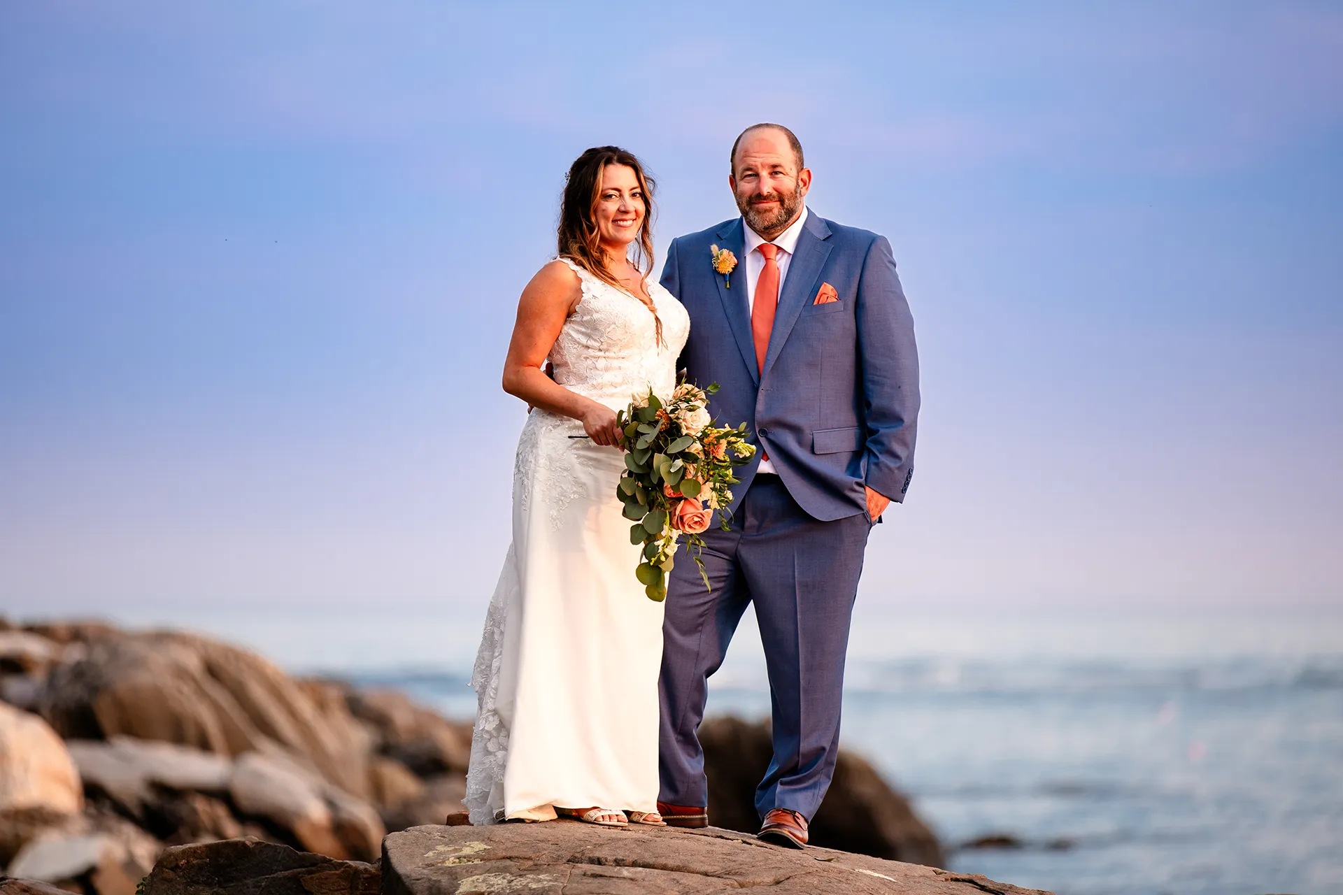 A newly married couple stand on a cliff and smile during their sunset wedding portraits at the Newagen Seaside Inn in Southport Maine.