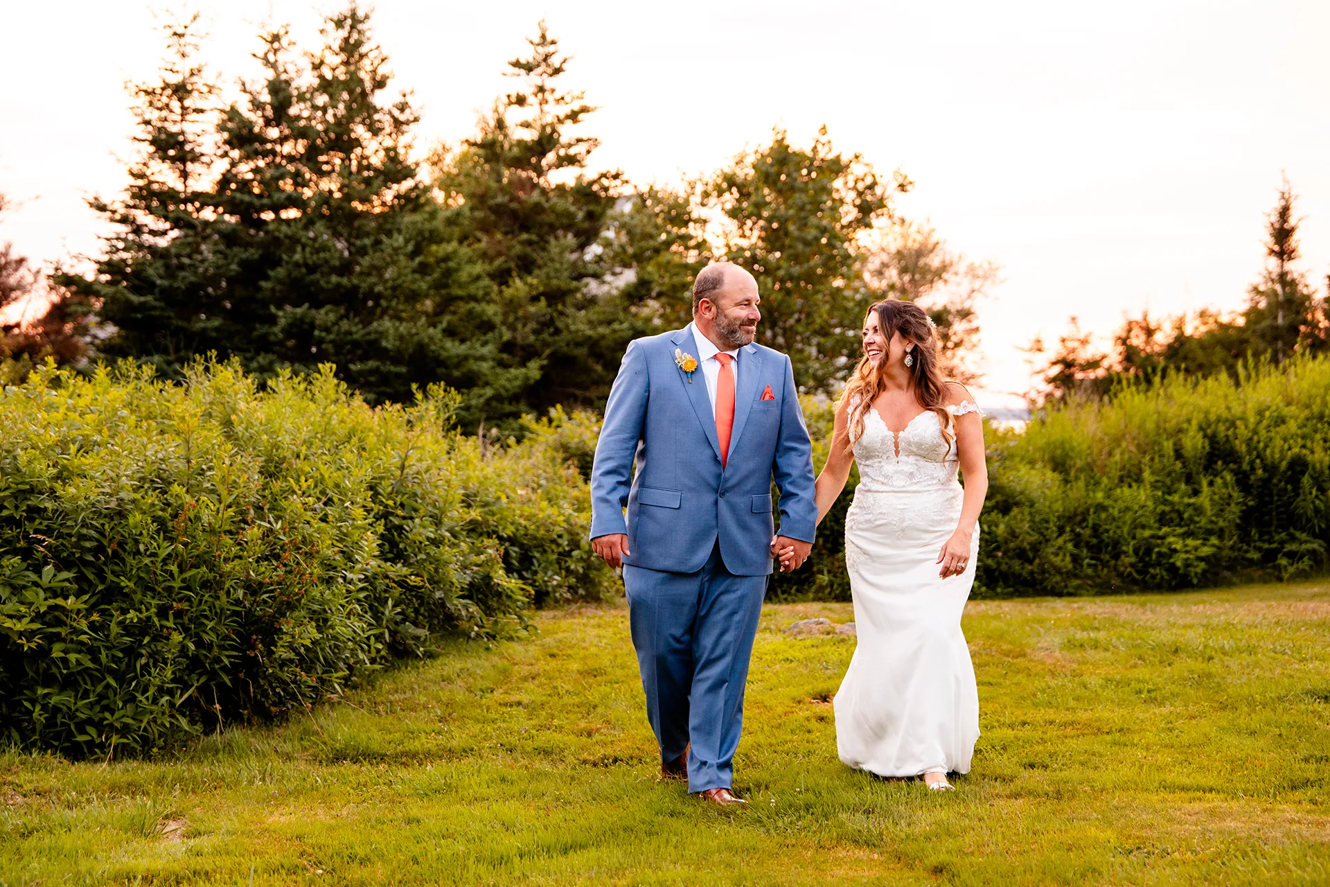 A bride and groom smile and hold hands as they walk through a field during wedding portraits at the Newagen Seaside Inn in Southport Maine.