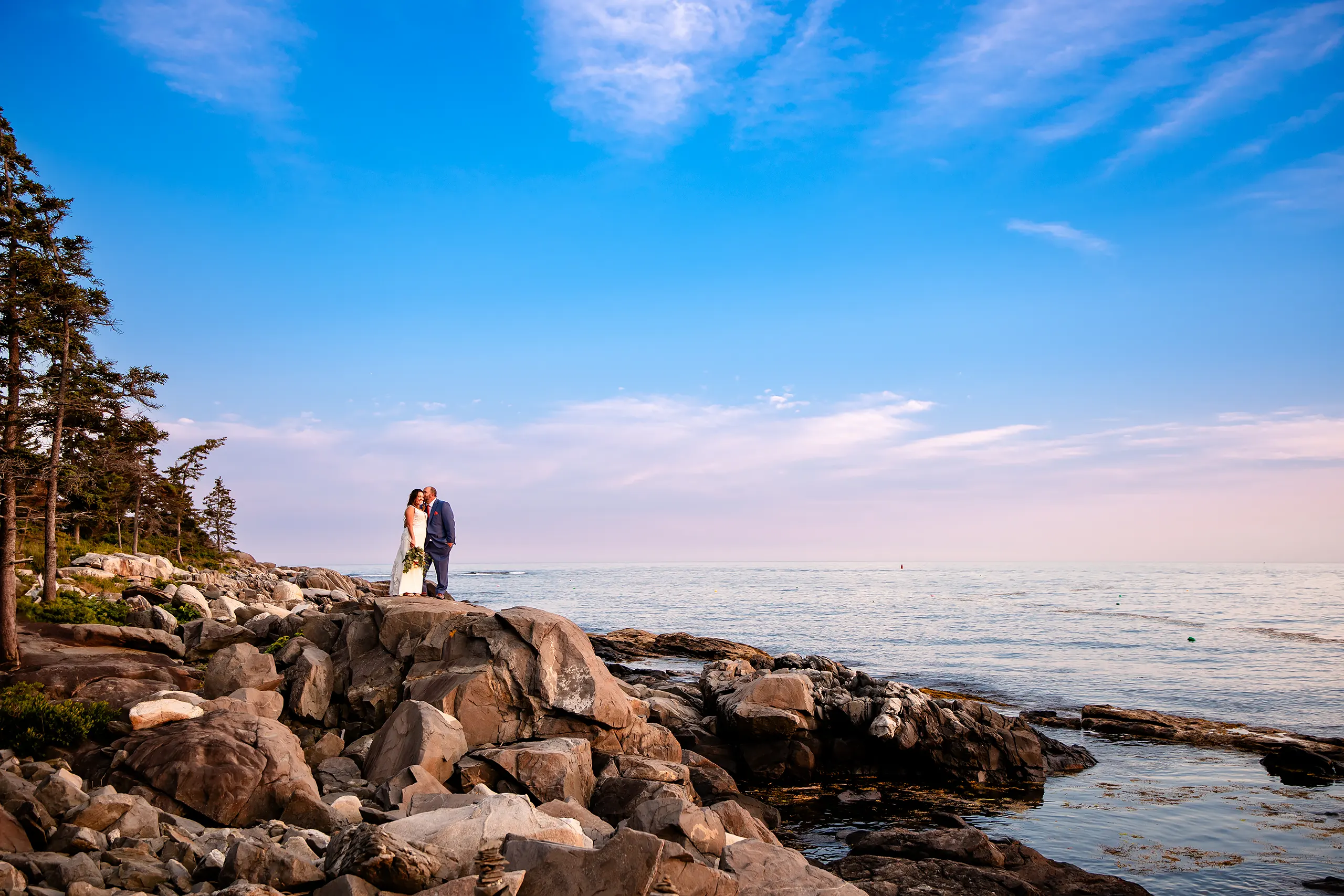 A groom kisses a bride on the cheek on a cliff at sunset during wedding portraits at the Newagen Seaside Inn in Southport Maine.