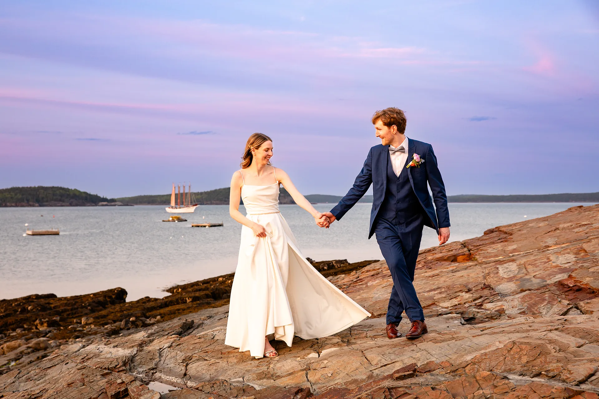 maine wedding photography caroline max 41 A bride and groom hold hands as they walk along a rocky coastline during wedding portraits in Bar Harbor, Maine.