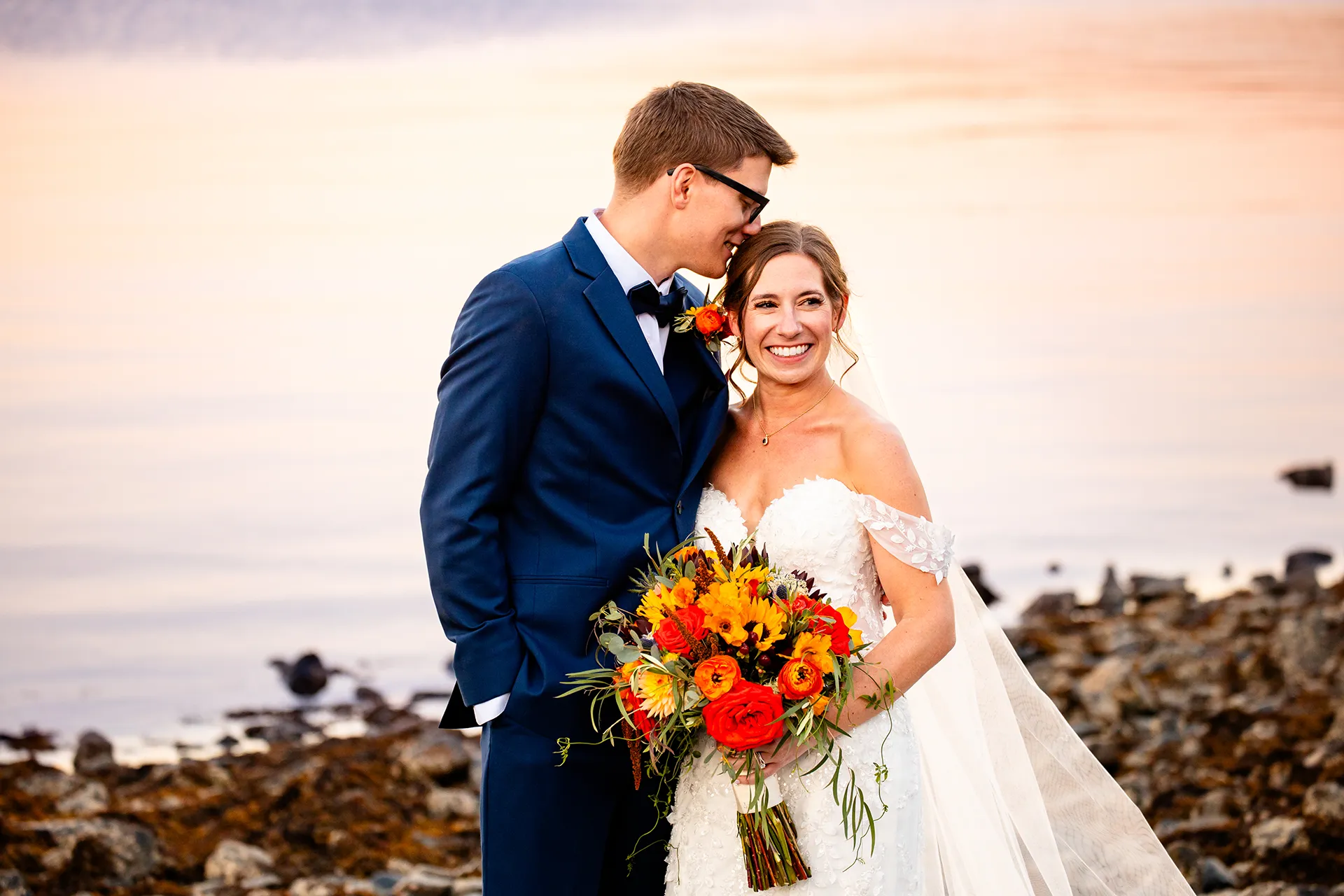 A groom snuggles into a bride during wedding portraits on a rocky beach at the Atlantic Oceanside Hotel & Event Center in Bar Harbor, Maine.