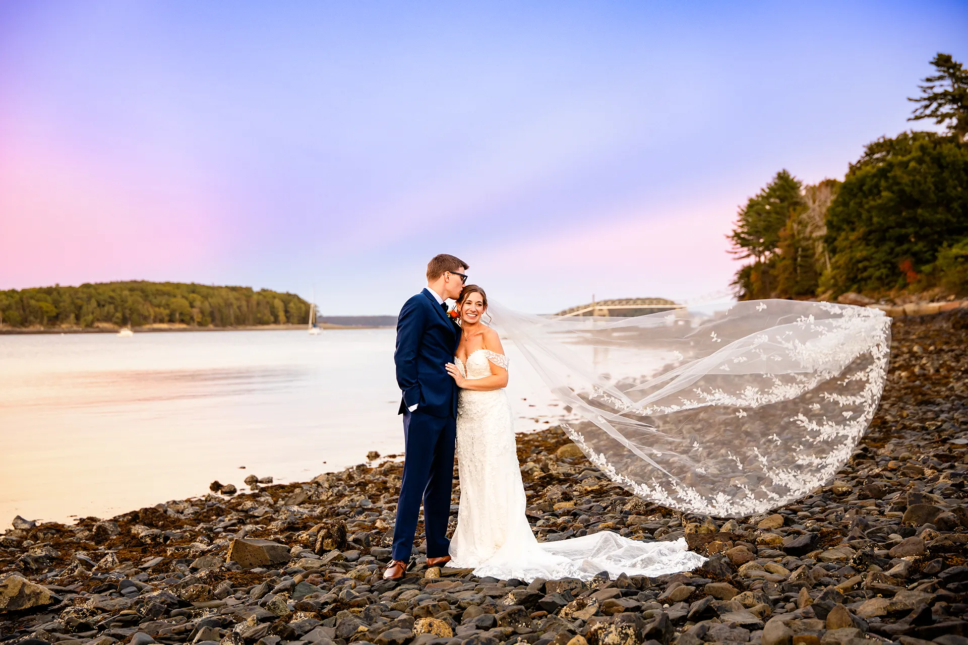 A groom kisses a bride on the head as her veil flies in the breeze on a beach at the Atlantic Oceanside Hotel & Event Center in Bar Harbor, Maine.