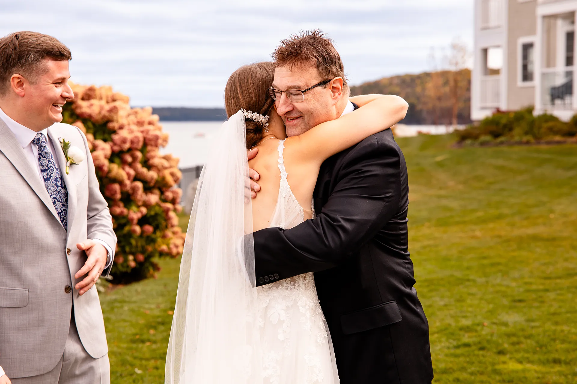 A dad hugs a bride as a groom laughs during a wedding ceremony at the Bar Harbor Inn and Spa near Acadia National Park in Maine.