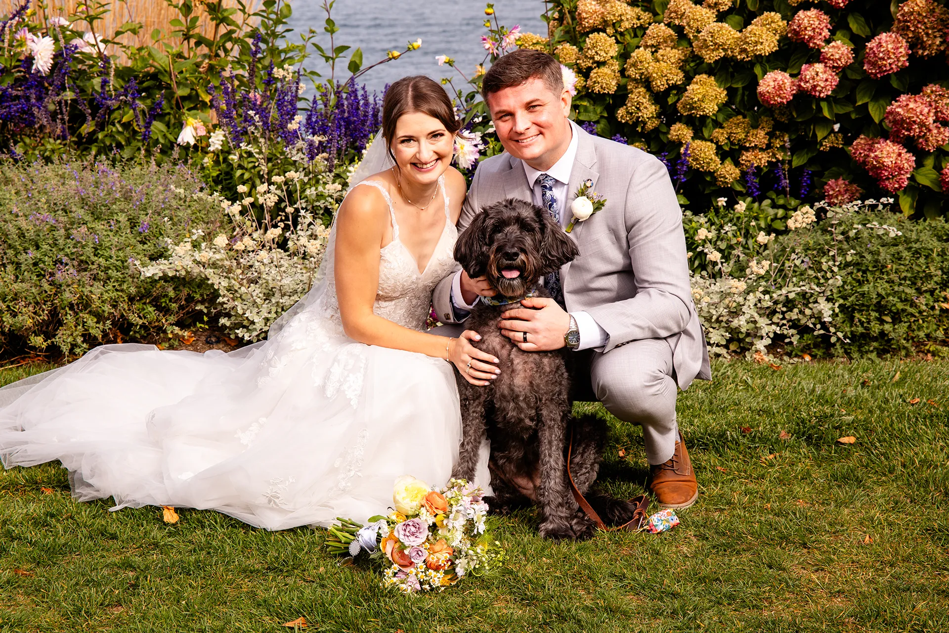 A bride and groom smile and pose with their Goldendoodle in front of flowers at the Bar Harbor Inn and Spa near Acadia National Park in Maine.