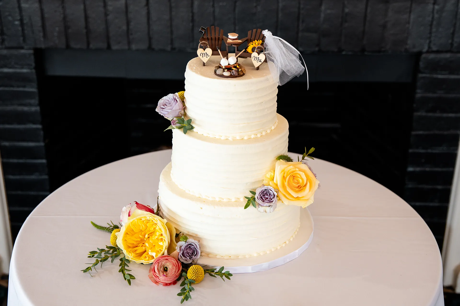 A white cake with a camp themed cake topper and flowers the the side for a wedding reception at the Bar Harbor Inn and Spa in Maine.