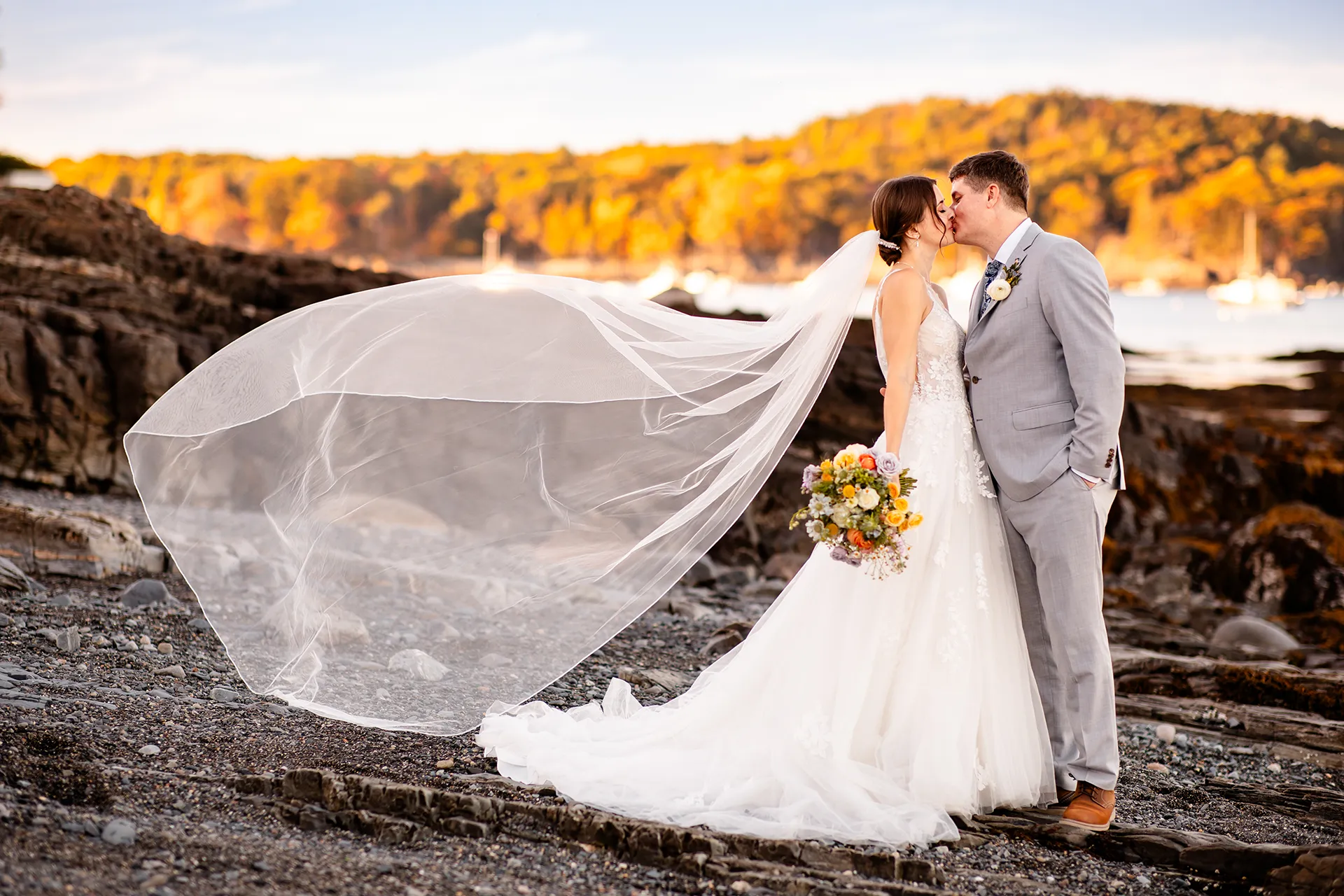 A bride's veil flies out behind her as she kisses a groom during wedding portraits on a rocky beach near the Bar Harbor Inn and Spa in Maine.
