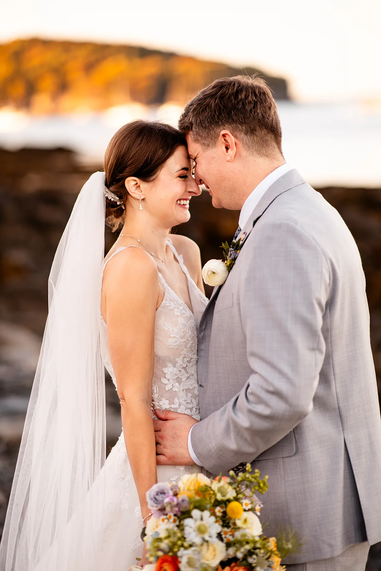 A bride and groom smile as they rest their heads together during wedding portraits on a rocky beach near the Bar Harbor Inn and Spa in Maine.