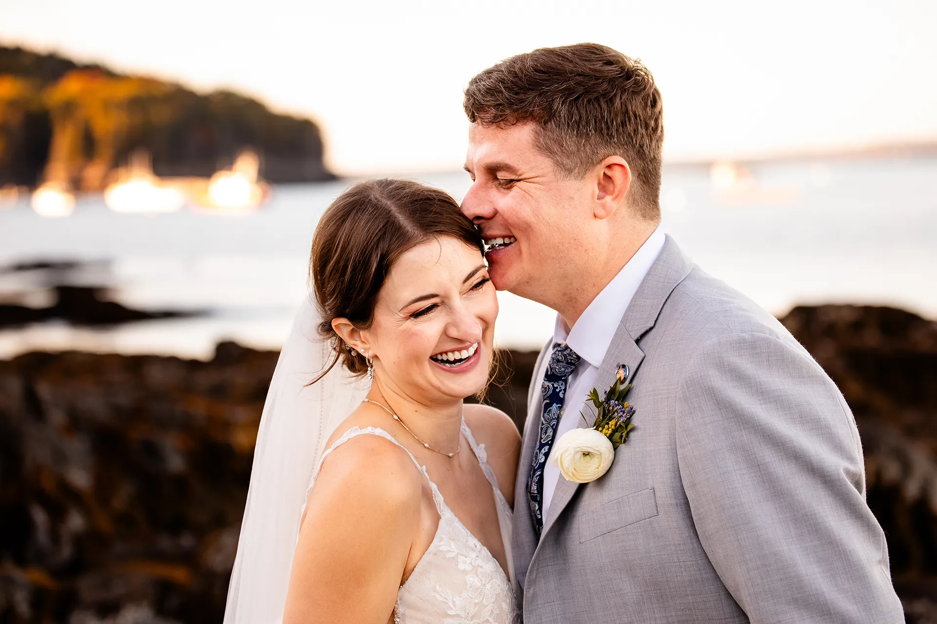 A newly married couple laugh as they snuggle together during wedding portraits on a rocky beach near the Bar Harbor Inn and Spa in Maine.