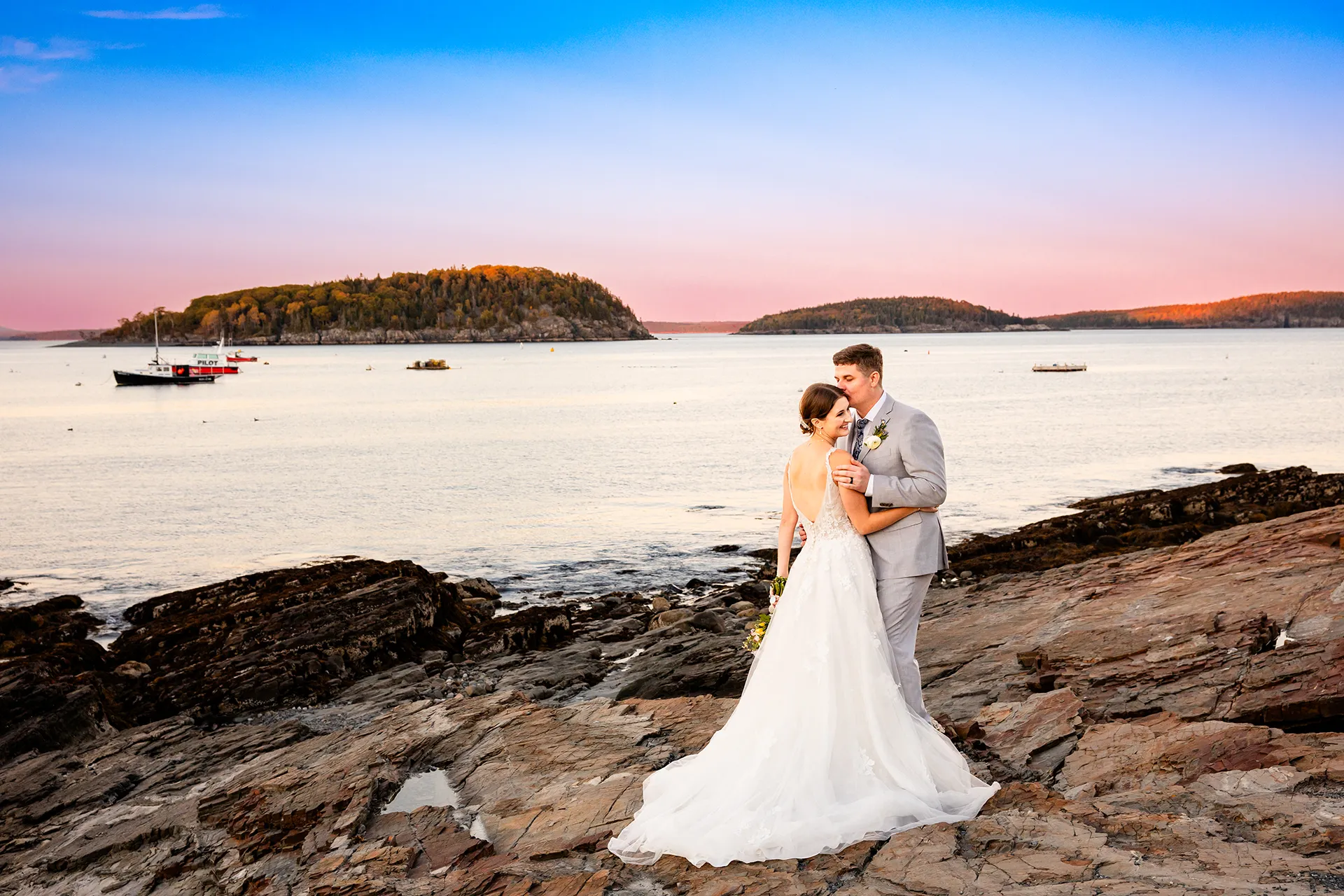 A bride smiles as a groom kisses her on the cheek during sunset wedding portraits at the Bar Harbor Inn near the Acadia National Park in Maine.