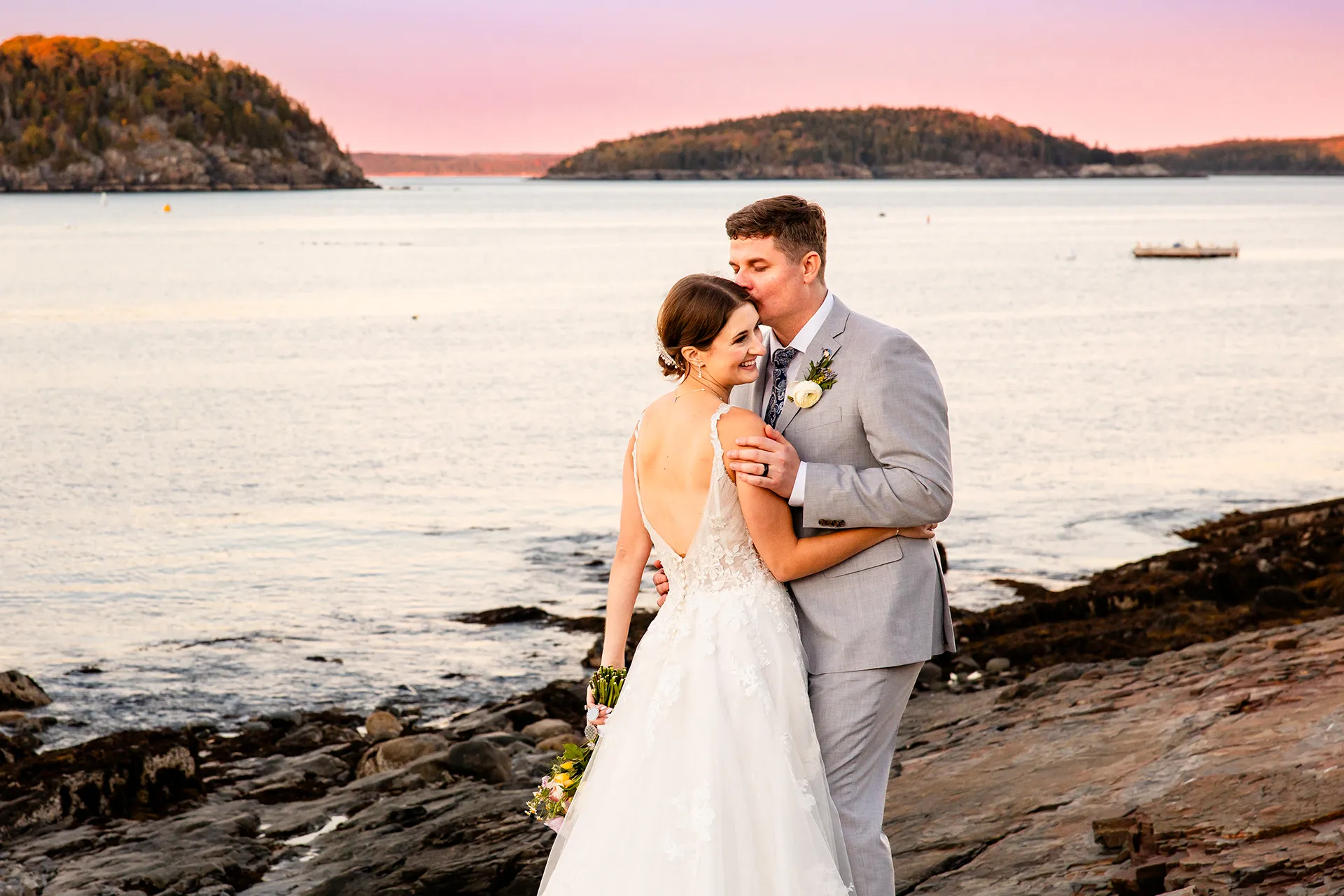 A bride smiles as a groom kisses her on the cheek in front of Frenchmans Bay on the property of the Bar Harbor Inn near the Acadia National Park in Maine.