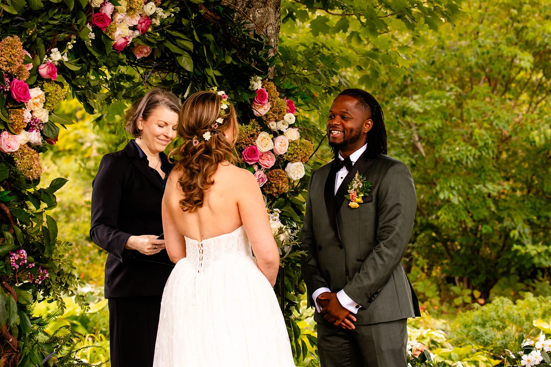 A groom smiles at a bride in front of a flower arbor during a wedding ceremony in Bangor, Maine.