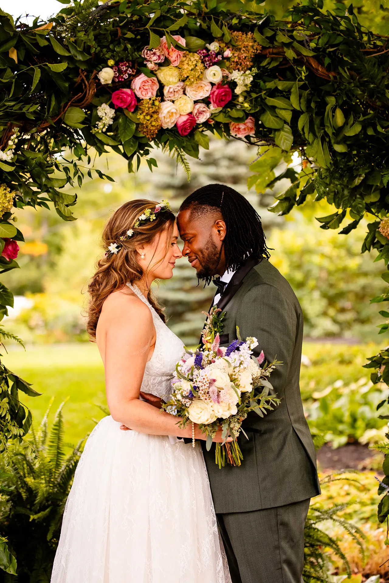 A recently married couple rest their heads together in front of a flower arbor during wedding portraits in Bangor, Maine.
