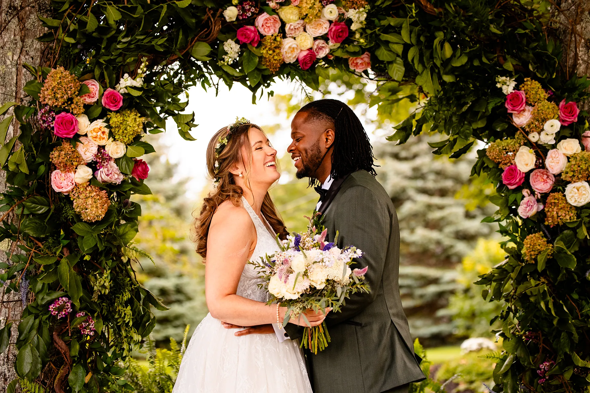 A bride and groom laugh as they pose in front of a flower arbor during wedding portraits in Bangor, Maine.